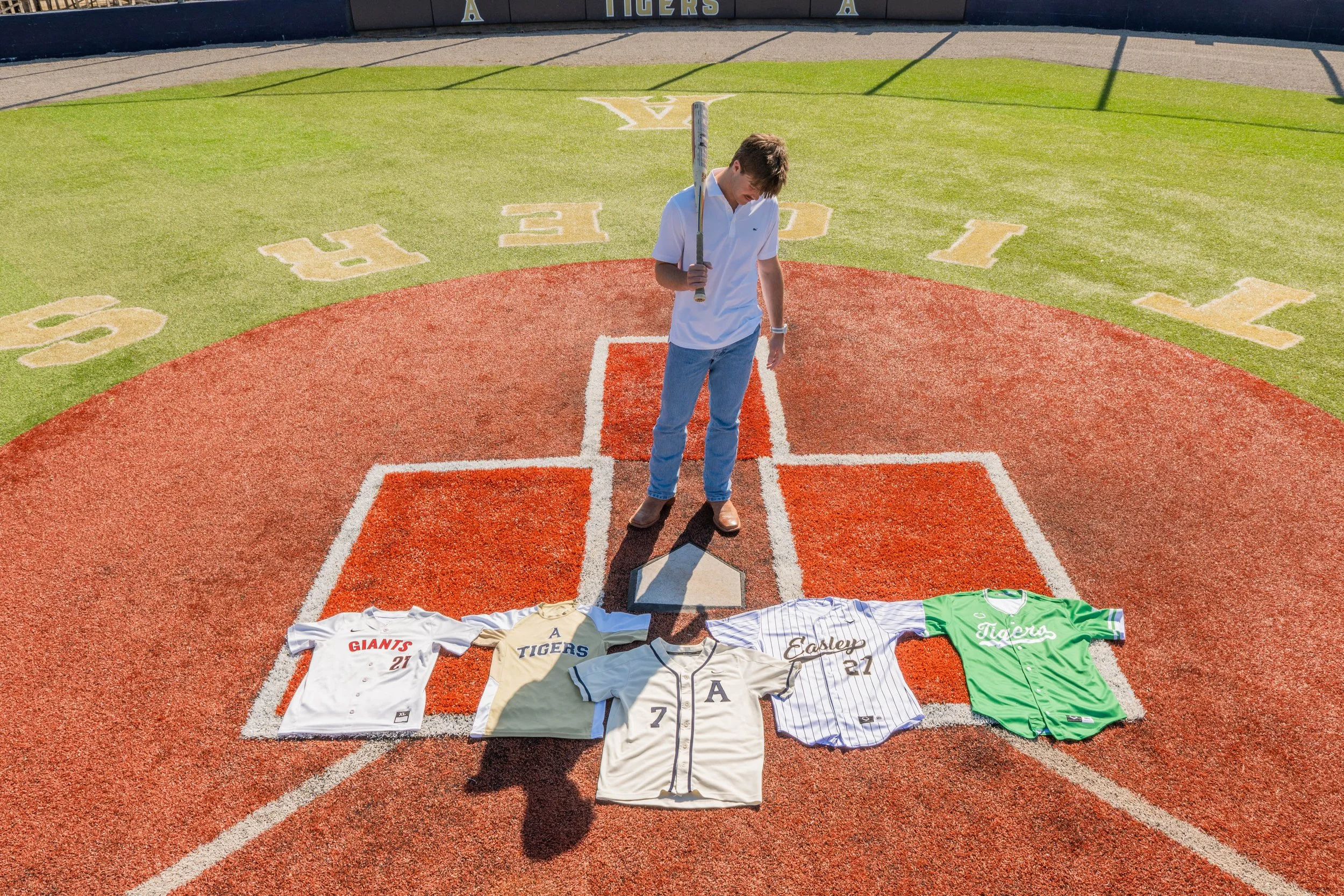 A young man standing on a baseball field holding a baseball bat, with baseball jerseys and shirts laid out on the ground in front of him, featuring team names and numbers, on a sunny day.