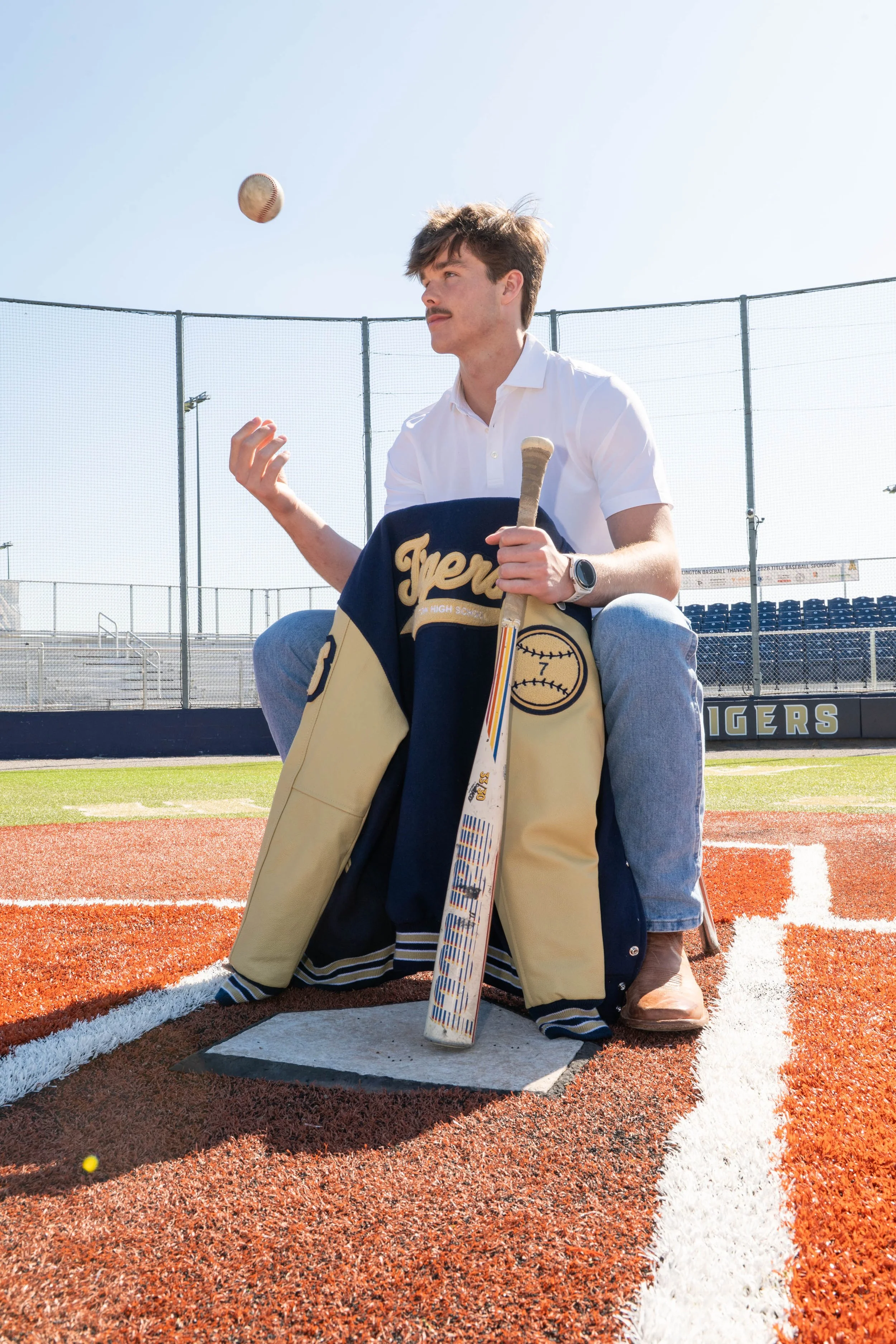 A young man kneeling on a sports field near a base marker, holding a baseball bat, with a baseball in the air above him.