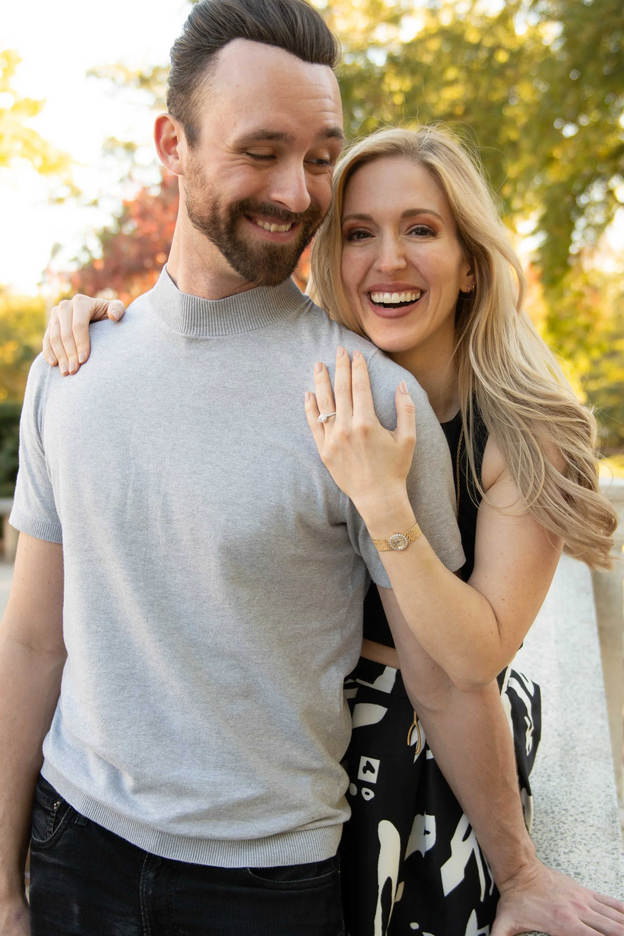 A happy couple outdoors, smiling and embracing. The man has dark hair and a beard, wearing a light gray shirt. The woman has long blonde hair, wearing a black dress with white abstract patterns, and a gold watch and ring. Autumn trees with yellow lea