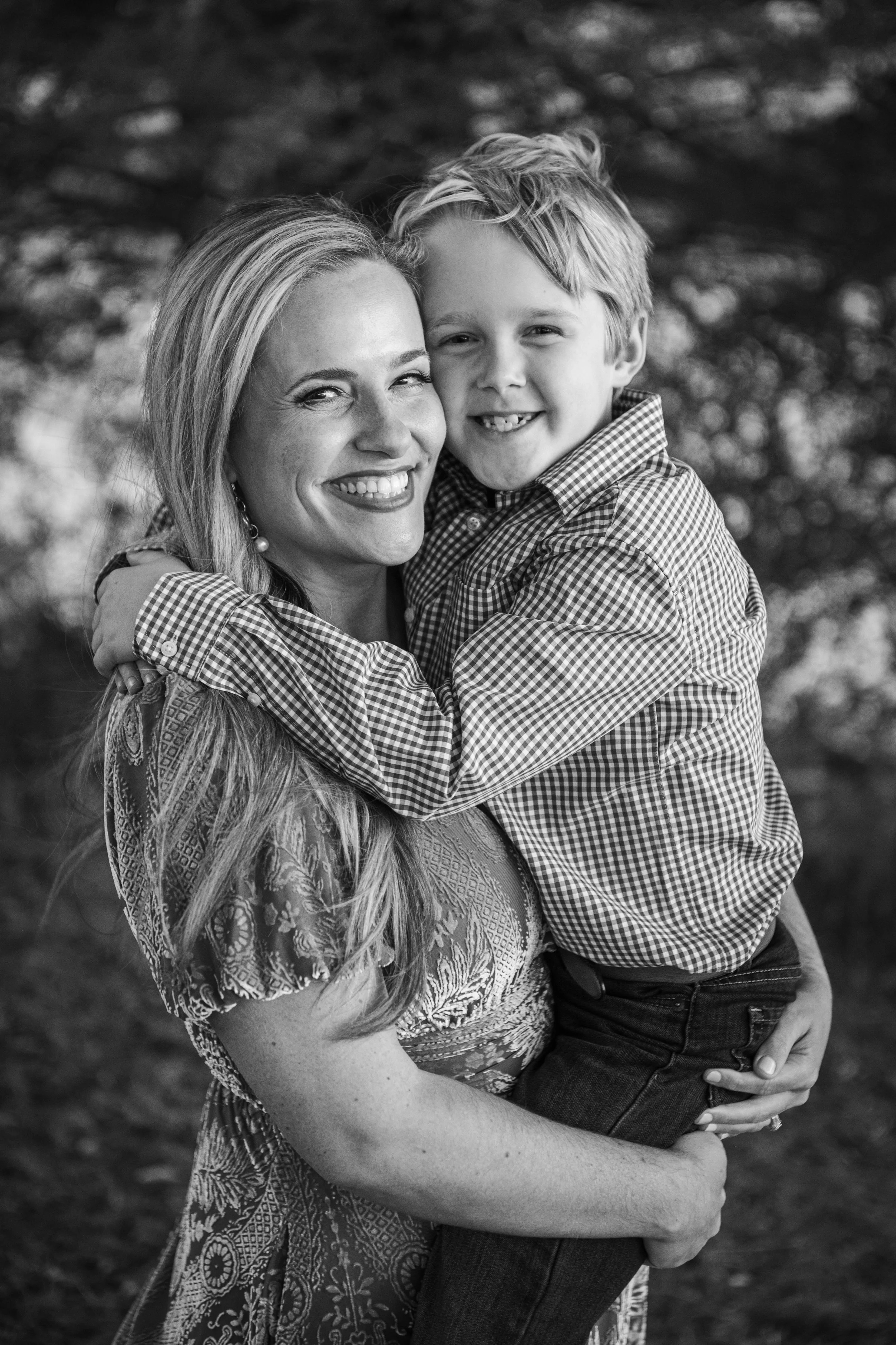 A woman with long, wavy hair and jewelry, and a young boy with curly hair, are hugging each other outdoors. Both are smiling and looking at the camera. The woman is wearing a patterned dress, and the boy is wearing a checkered shirt.