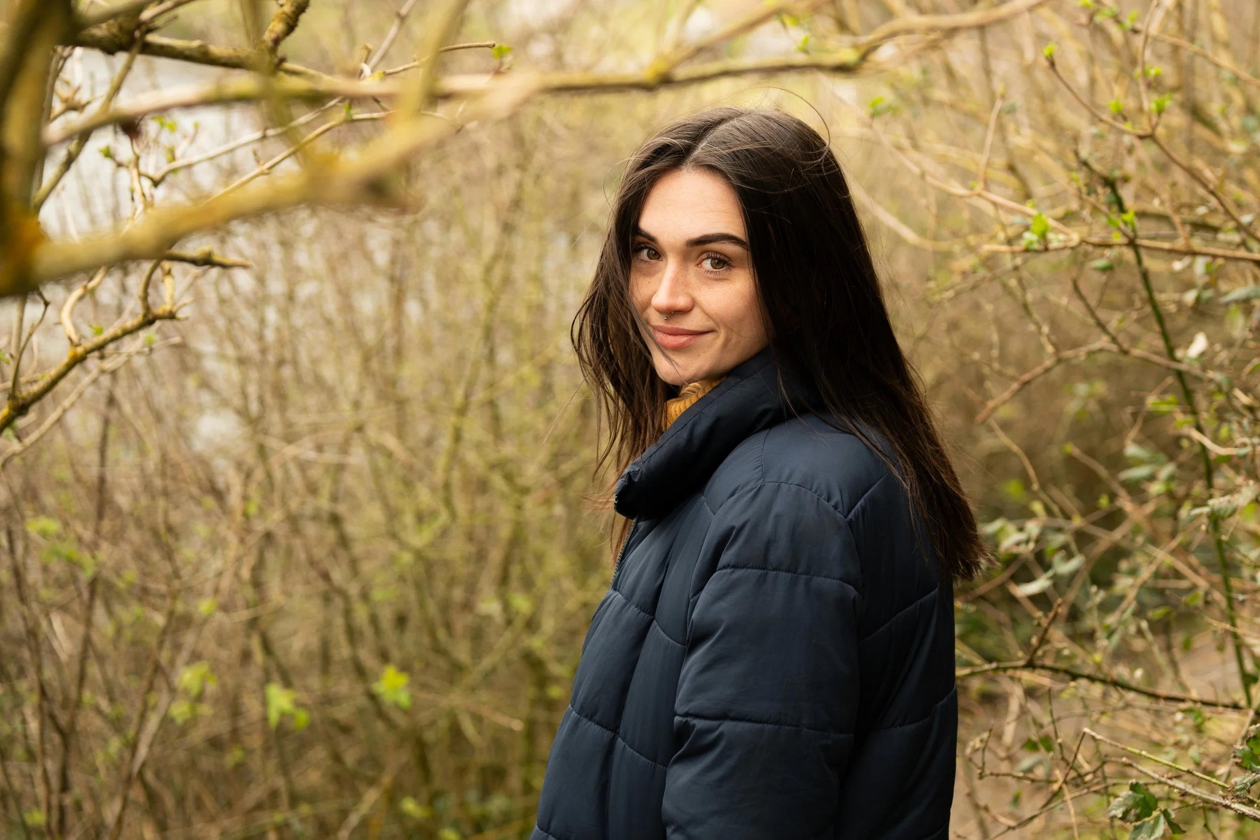 A young woman with long dark hair and a septum piercing stands outdoors among leafless bushes, smiling softly at the camera.