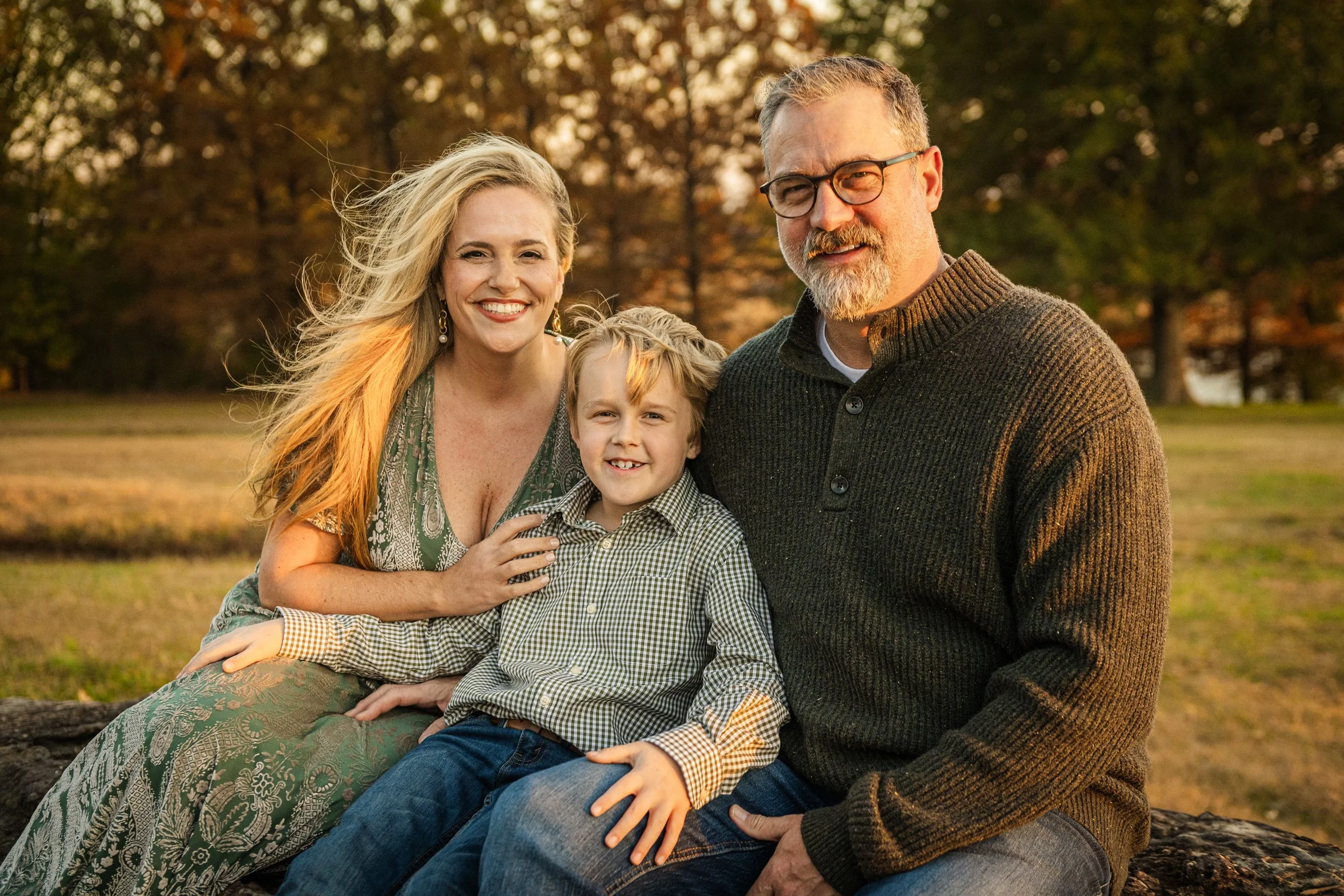 A family of three, a woman, a man, and a young boy, sitting outdoors on a log with trees in the background during fall, smiling at the camera.