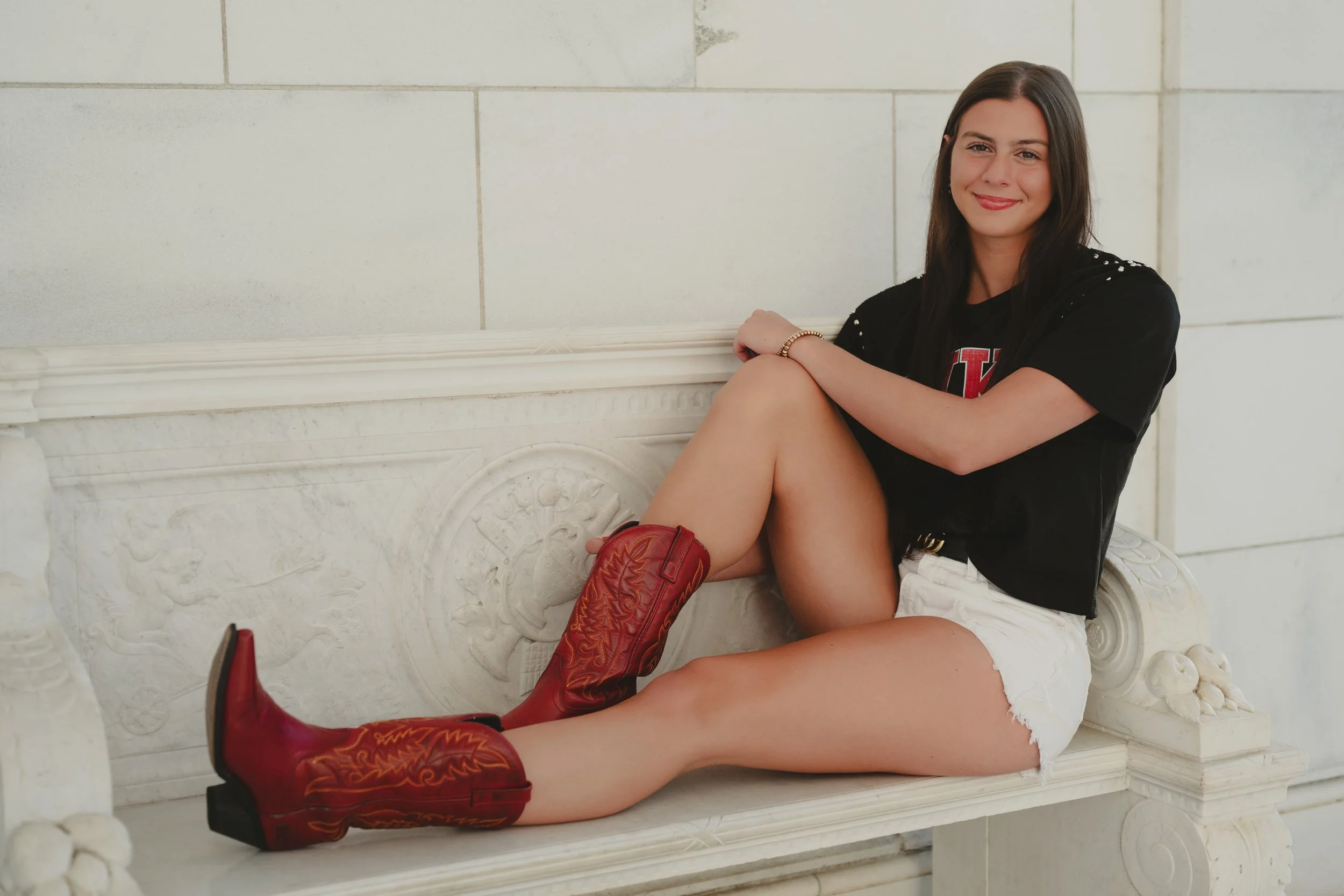 A young woman with long dark hair smiling, sitting on a decorative white marble bench in front of a white wall, wearing a black T-shirt, white shorts, and red cowboy boots.