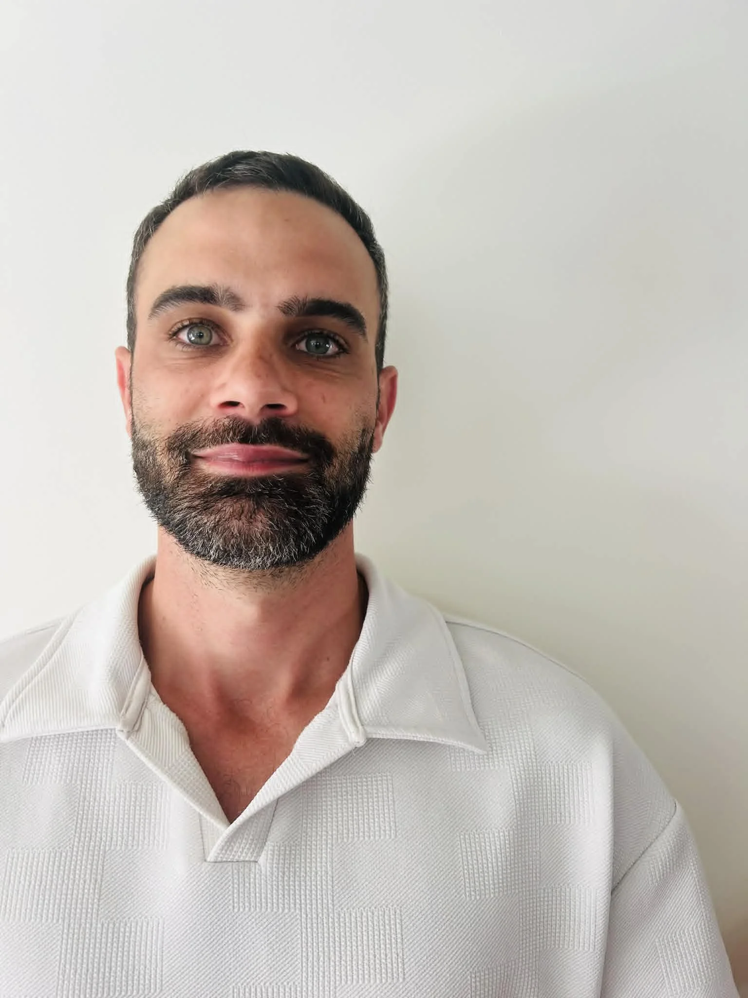 Close-up of a man with dark hair, a beard, and hazel eyes, wearing a white collared shirt, standing against a plain off-white wall.