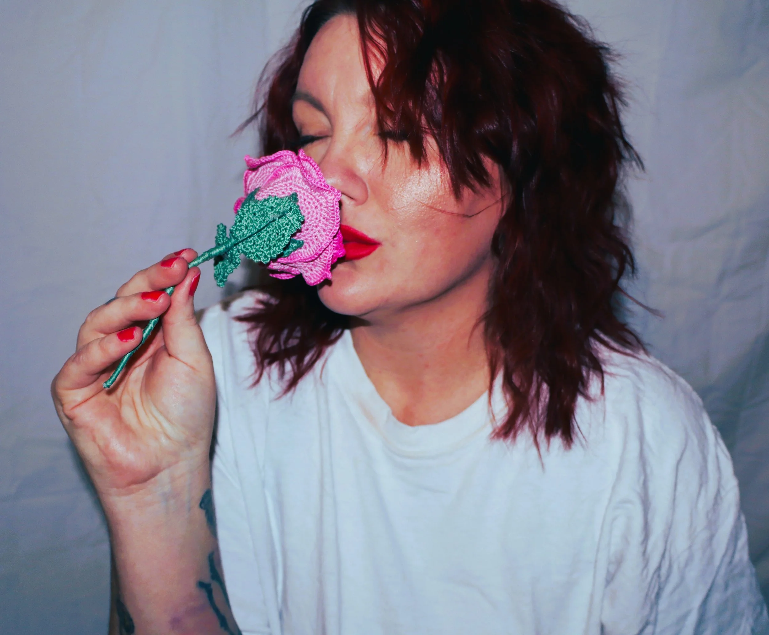 A woman with red curly hair and red lipstick holding a pink and green crocheted flower close to her face, eyes closed.