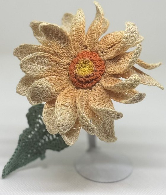 A crocheted flower with petals in light beige and a yellow and orange center, along with a green crocheted stem and leaf.
