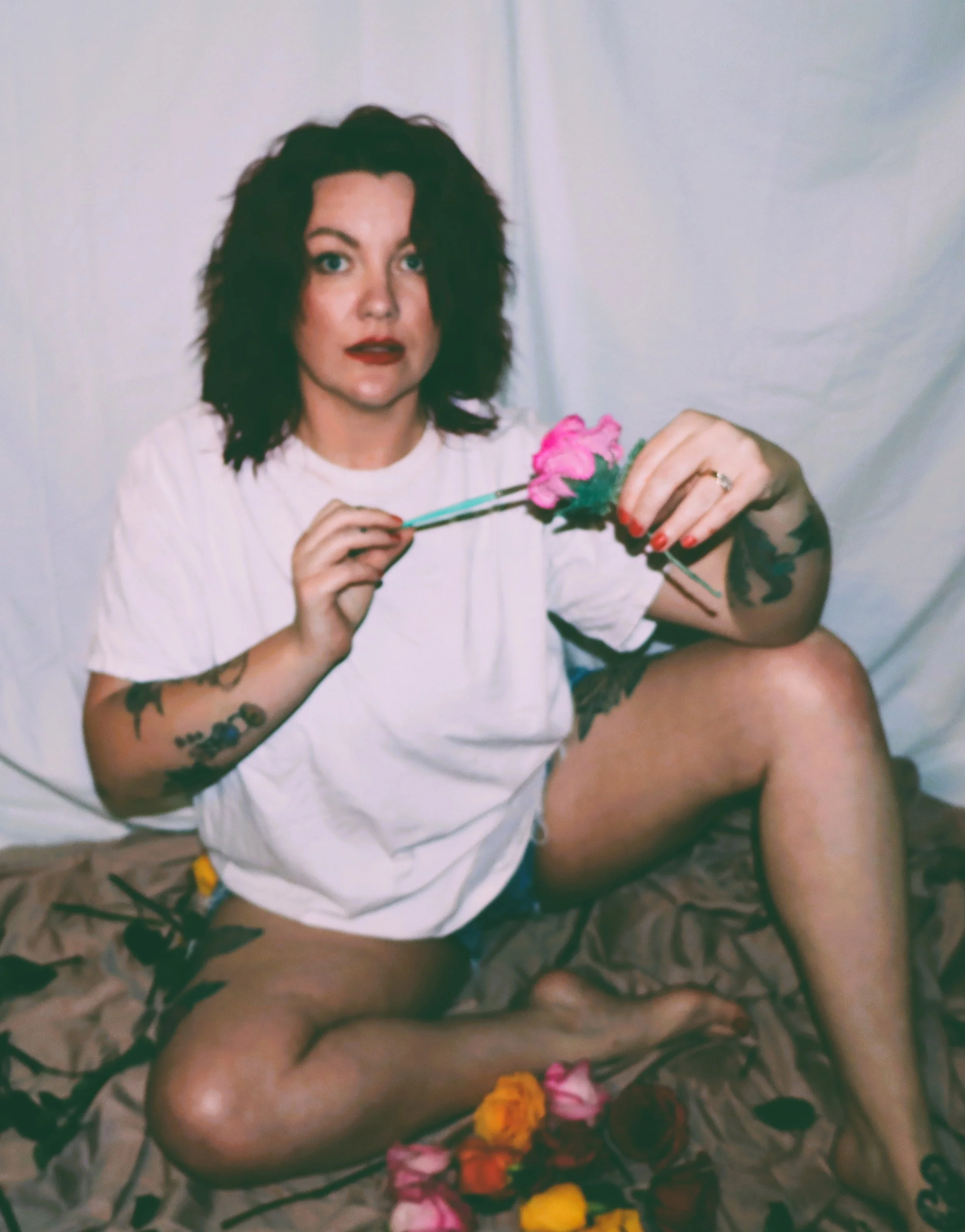 A woman with dark hair and tattoos on her arms holding a pink flower with a background of scattered flowers on the ground.