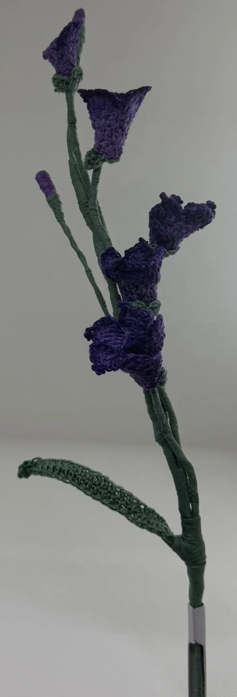 A handmade flower embroidery with purple and blue petals and green leaves on a gray background.
