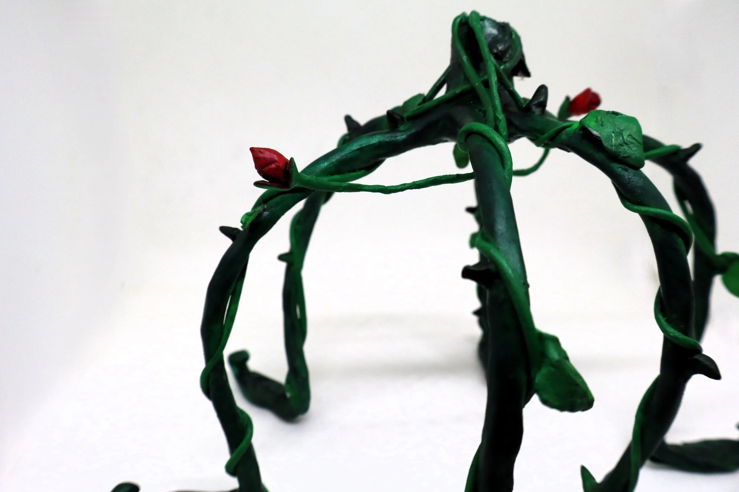 A close-up of a green wire crown with small red and black plastic buds on a plain white background.