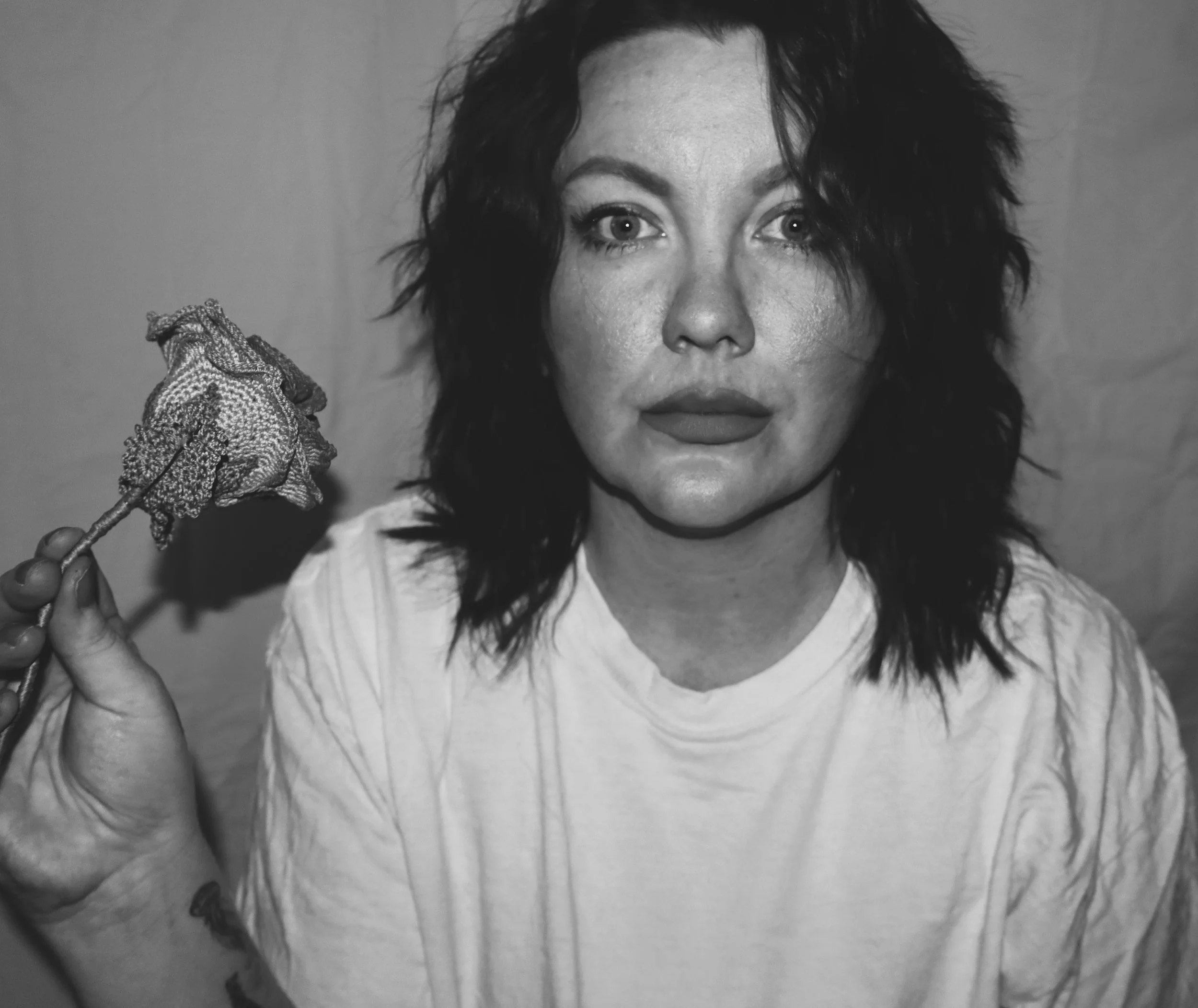 Black and white photo of a woman with curly hair, looking directly at the camera with a slight smile. She is holding a dried flower with textured petals close to her face.