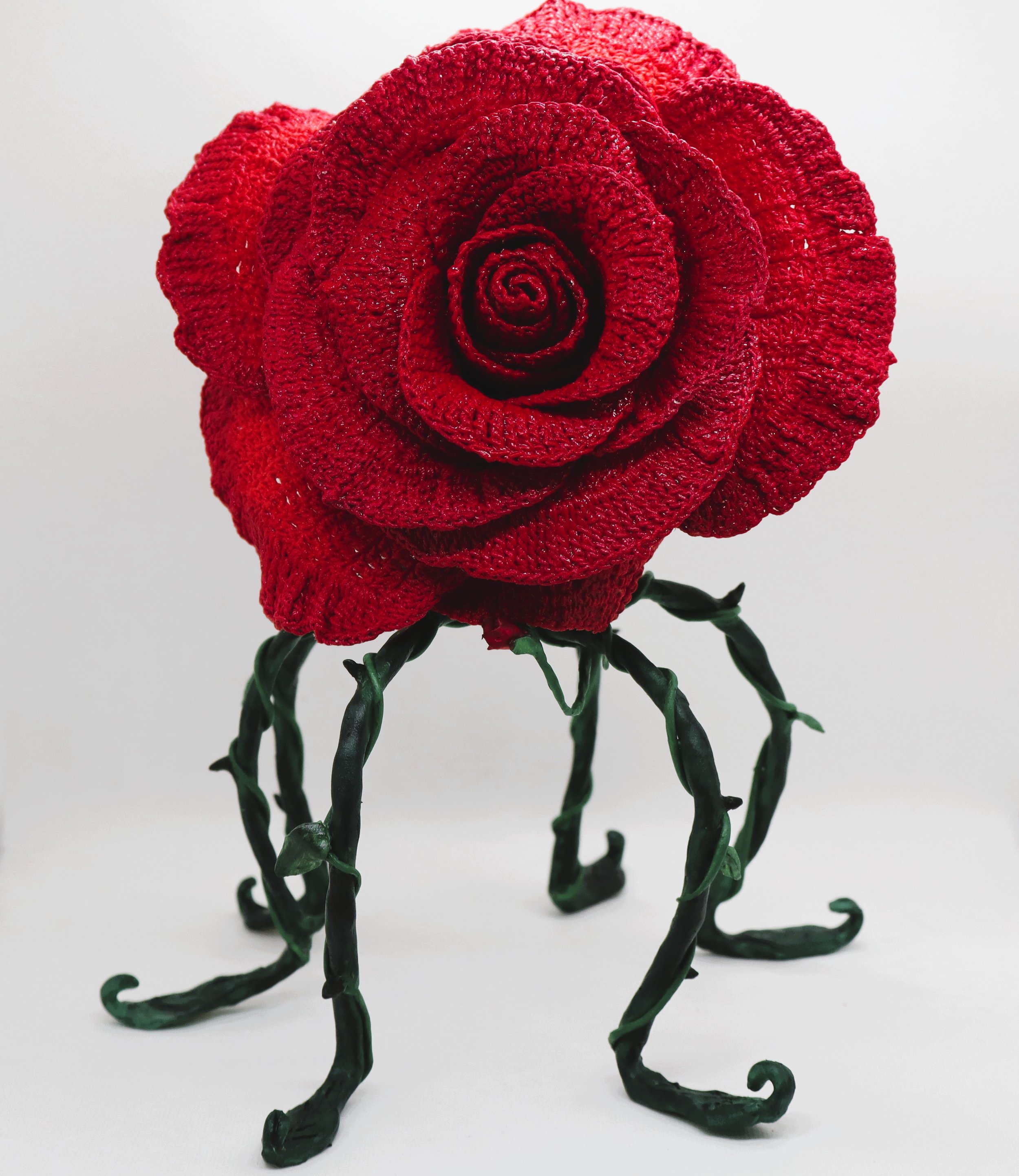 A metal sculpture of a red rose with curly green stem and leaves, set against a plain white background.