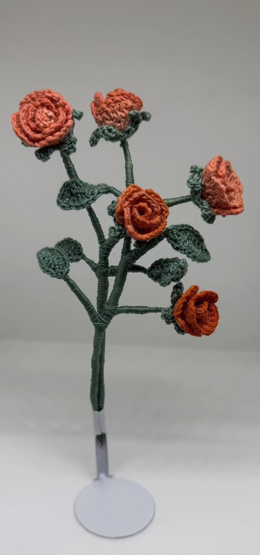 Handcrafted wire and yarn flower bouquet with orange knitted roses and green wire stems and leaves on a display stand.