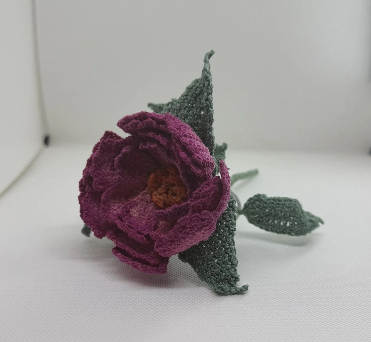 A crocheted pink rose with green leaves on a white surface.