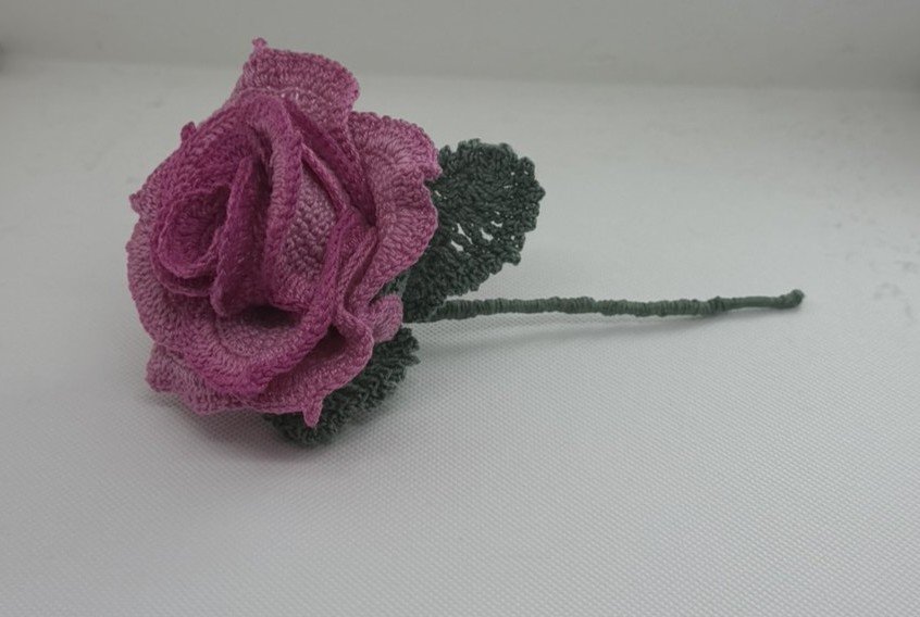 A pink, fabric flower with green leaves and stem placed on a white surface.