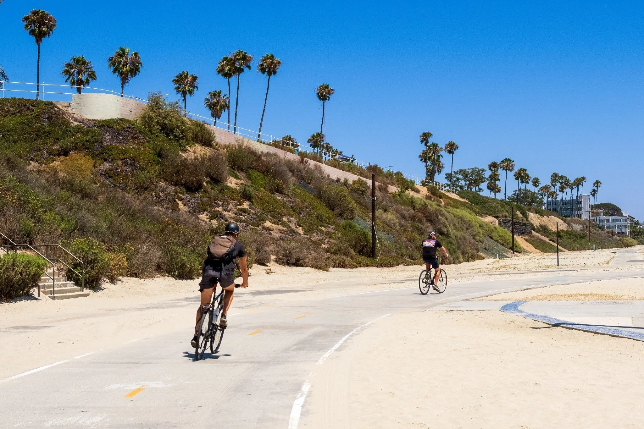 Two people riding bicycles on a paved path along a beach with sand and hills covered in bushes and palm trees, under a clear blue sky.