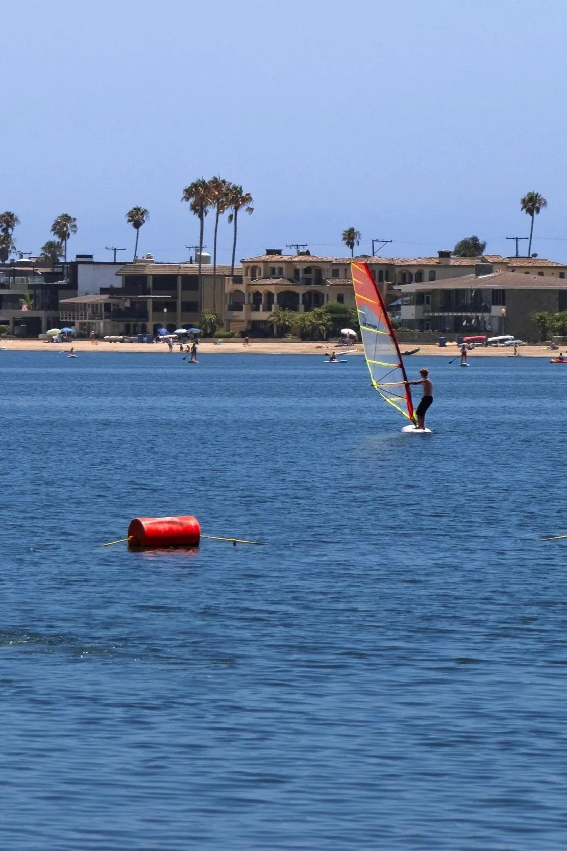 Person windsurfing on calm blue water near a sandy beach with houses and palm trees in the background.