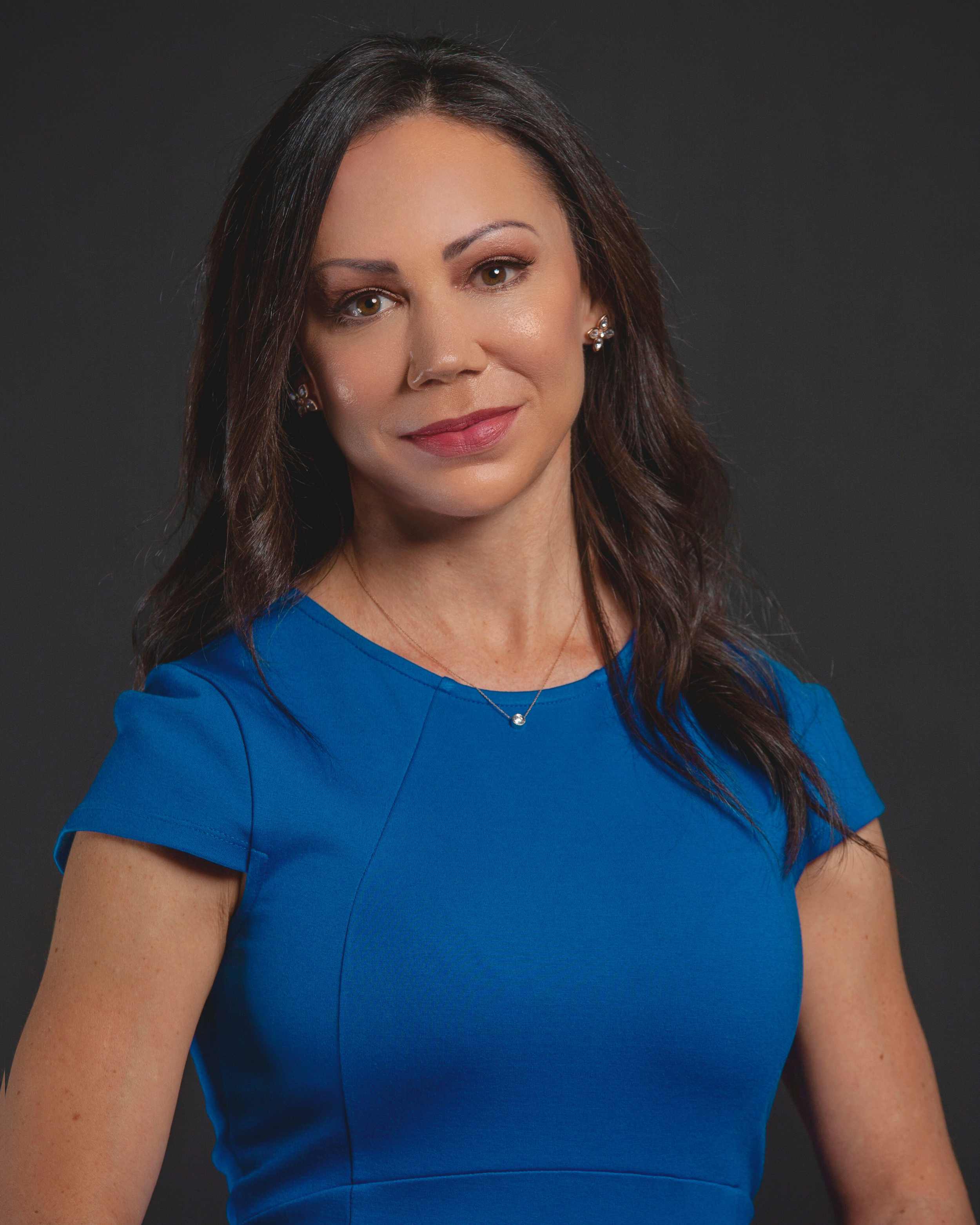 Close-up of a woman with brown hair and light skin, wearing a blue top and silver earrings, smiling softly against a dark background.