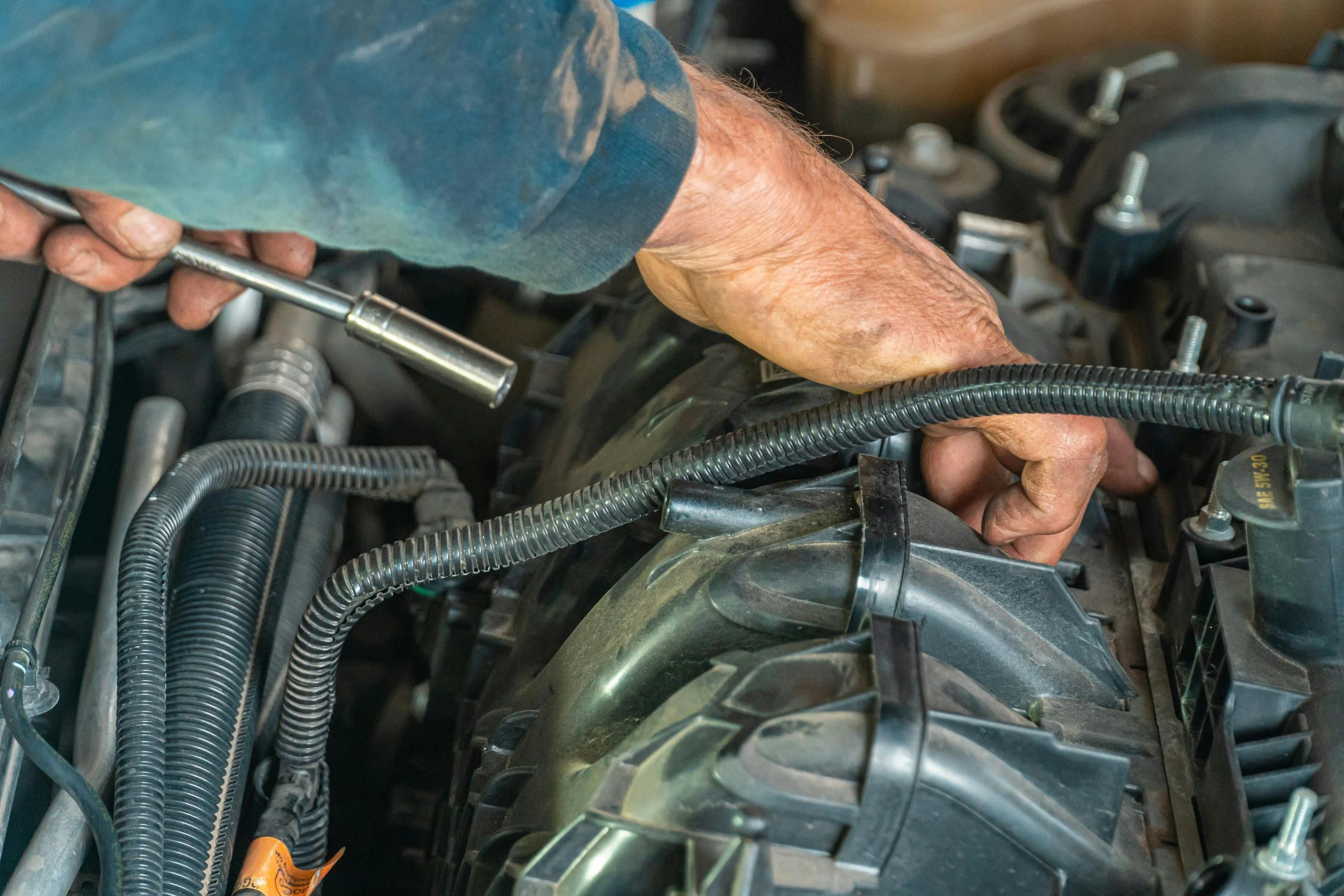 A person's hand working on a vehicle's engine using a wrench amidst hoses and engine components.