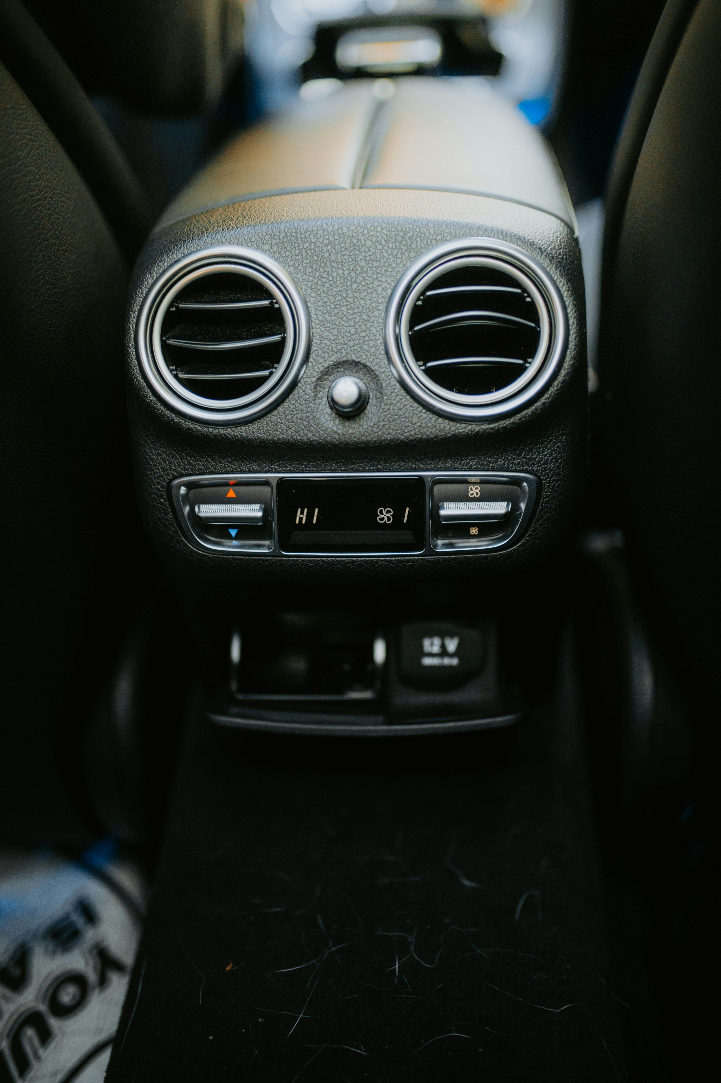The image shows the back of a car's center console with two circular air vents and climate control buttons beneath them. A portion of the passenger's shoe is visible in the lower left corner, and some loose hair or threads are scattered on the dark floor.