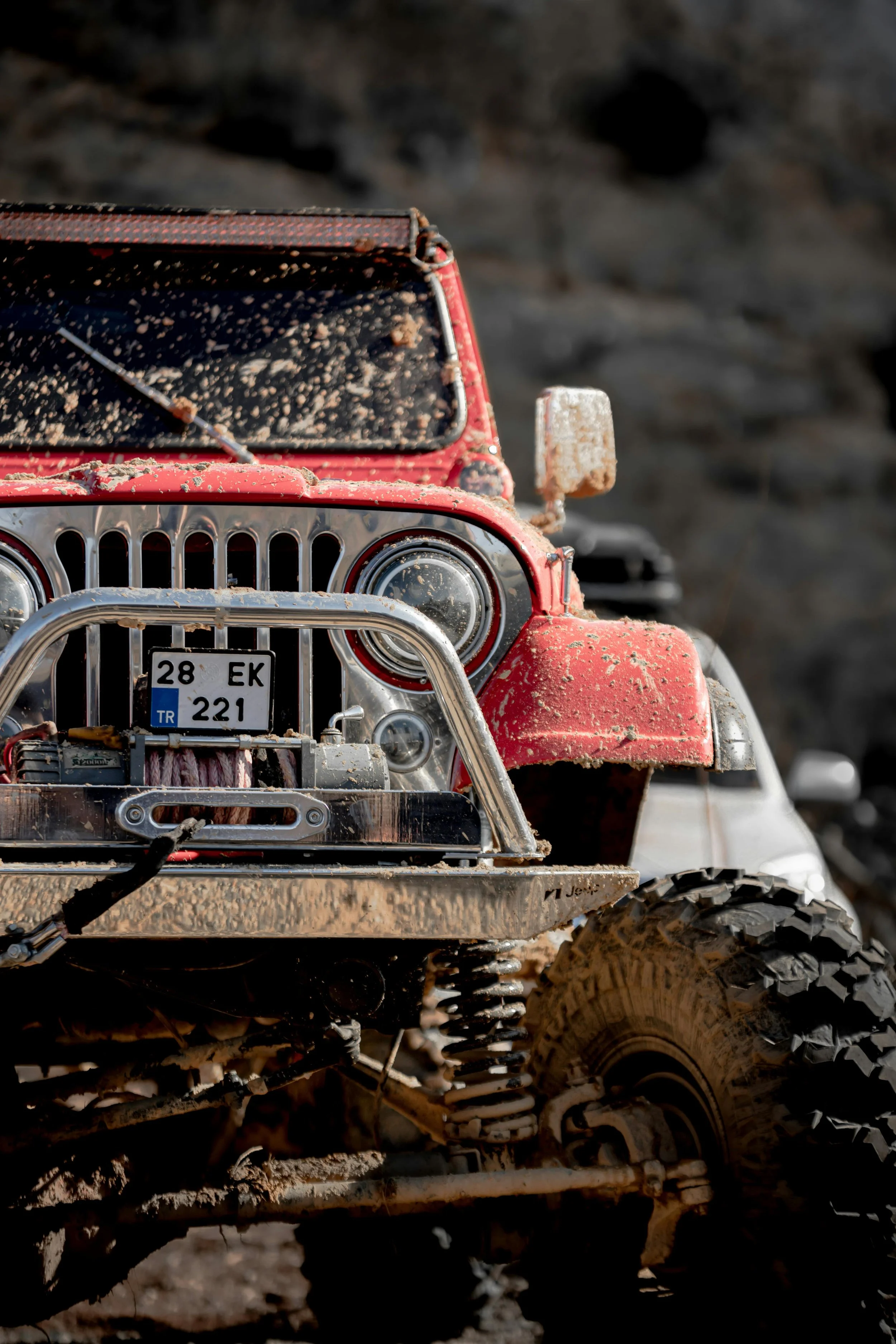 Red off-road vehicle covered in mud, with large rugged tires and a front bumper guard, partially visible in a muddy outdoor environment.
