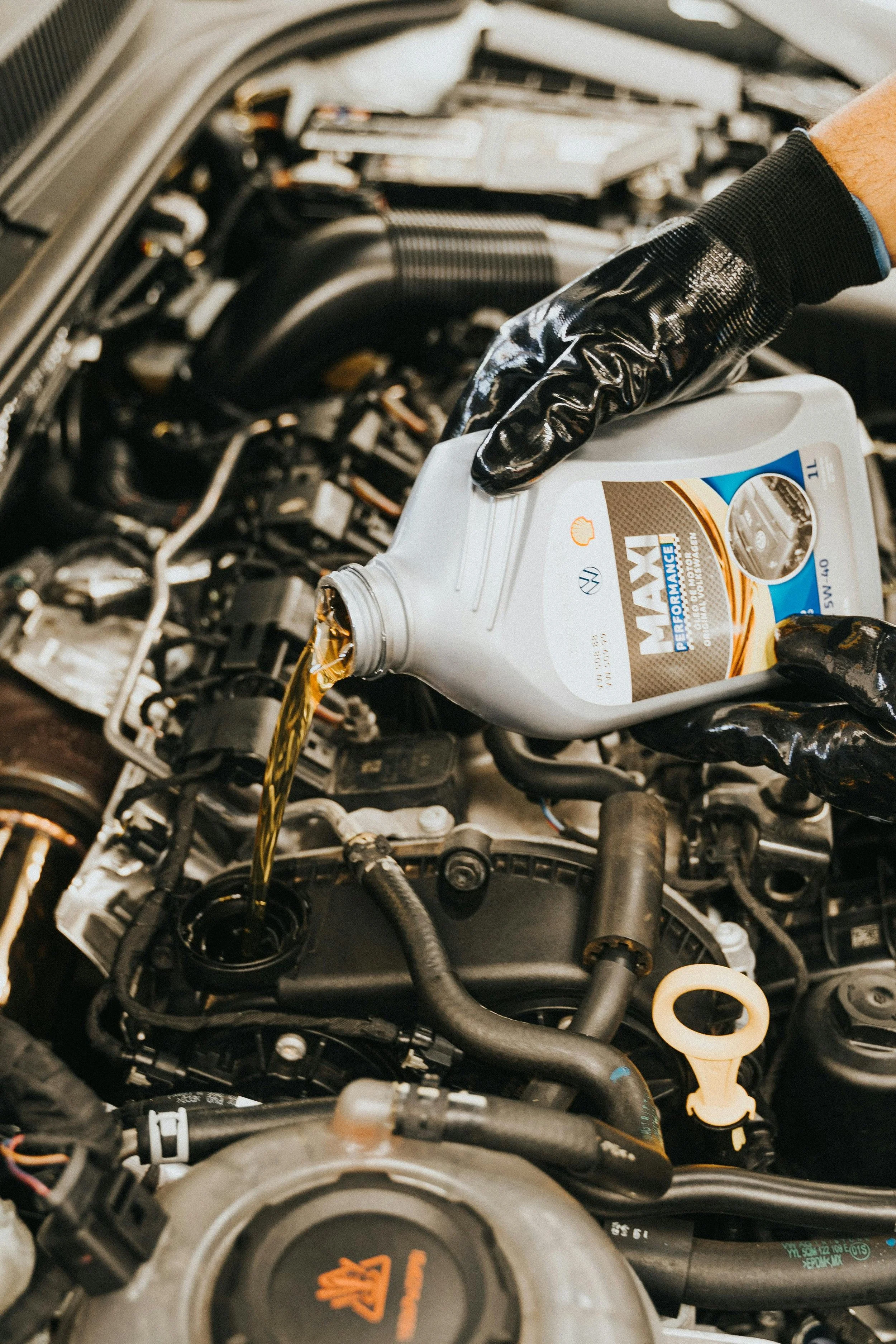 A person wearing black gloves pouring motor oil from a white container into the engine of a vehicle.