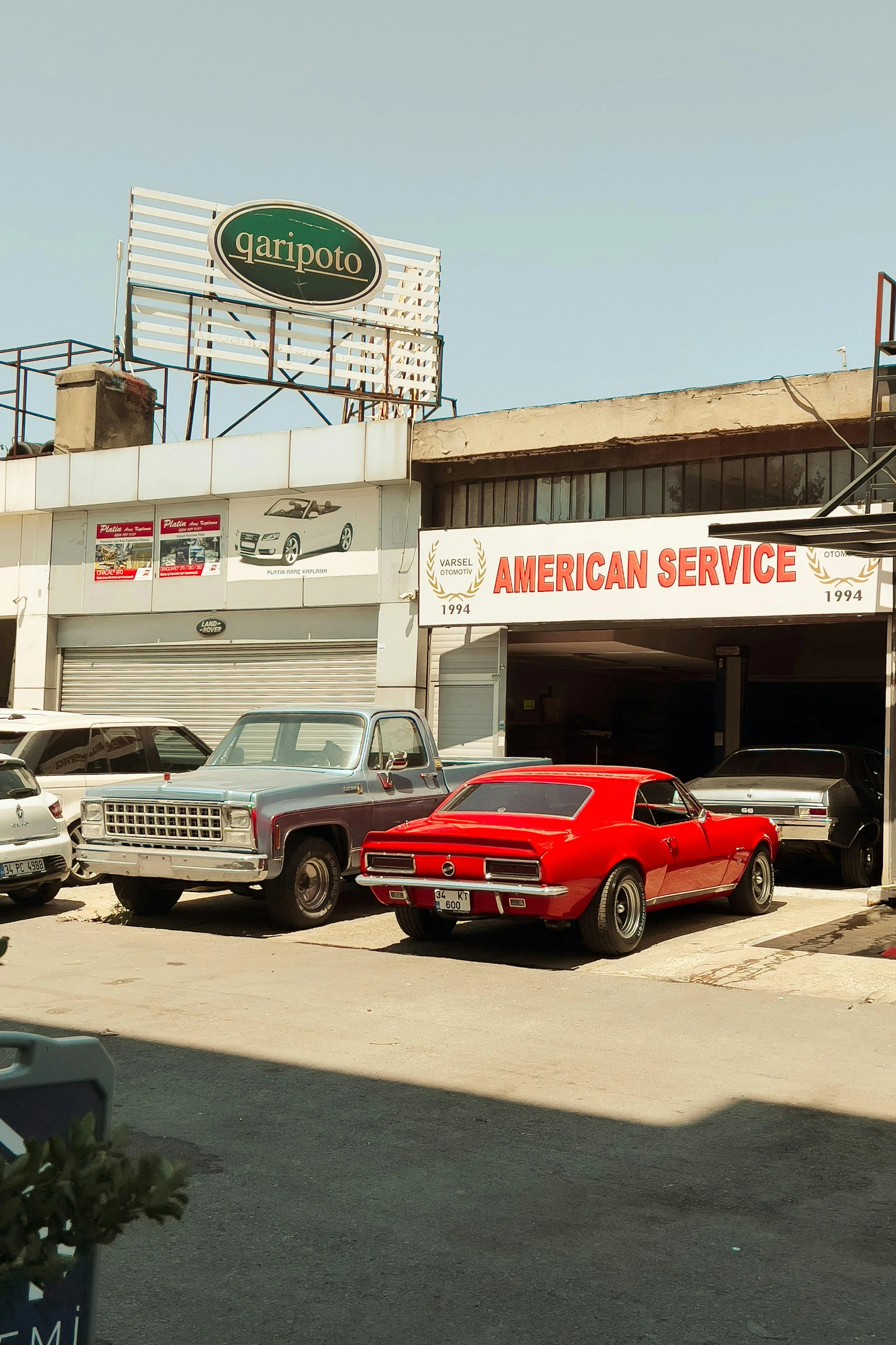 A parking lot with several vintage and modern cars parked in front of a building that has a sign reading 'American Service' and pictures of cars displayed on the wall. The building has a sign with the words 'Garipoto' on the roof.