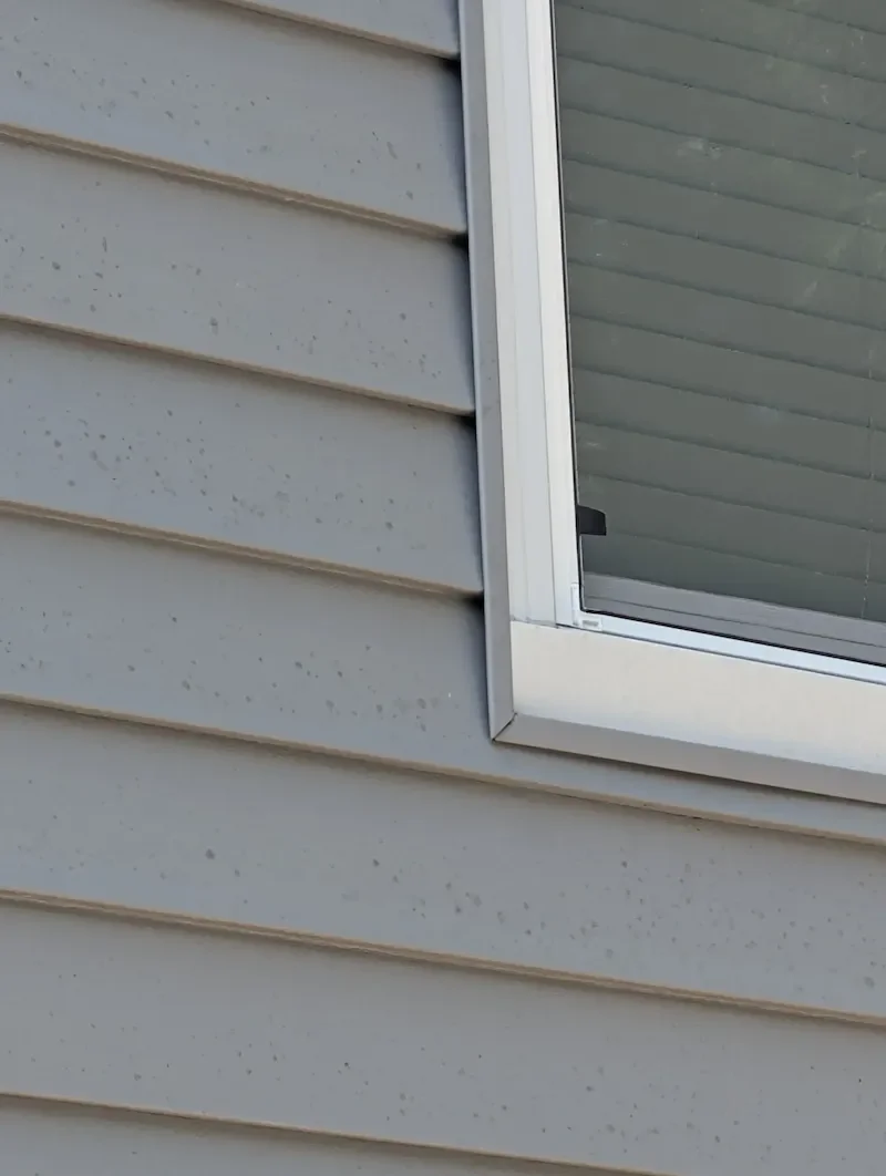 Close-up of vinyl siding gap at window corner showing how high-pressure washing forces water behind panels creating hidden mold and moisture damage