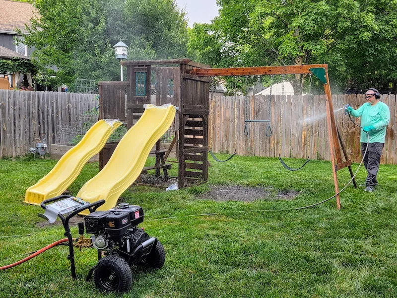 Professional applying low-pressure cleaning solution to backyard playset with full PPE