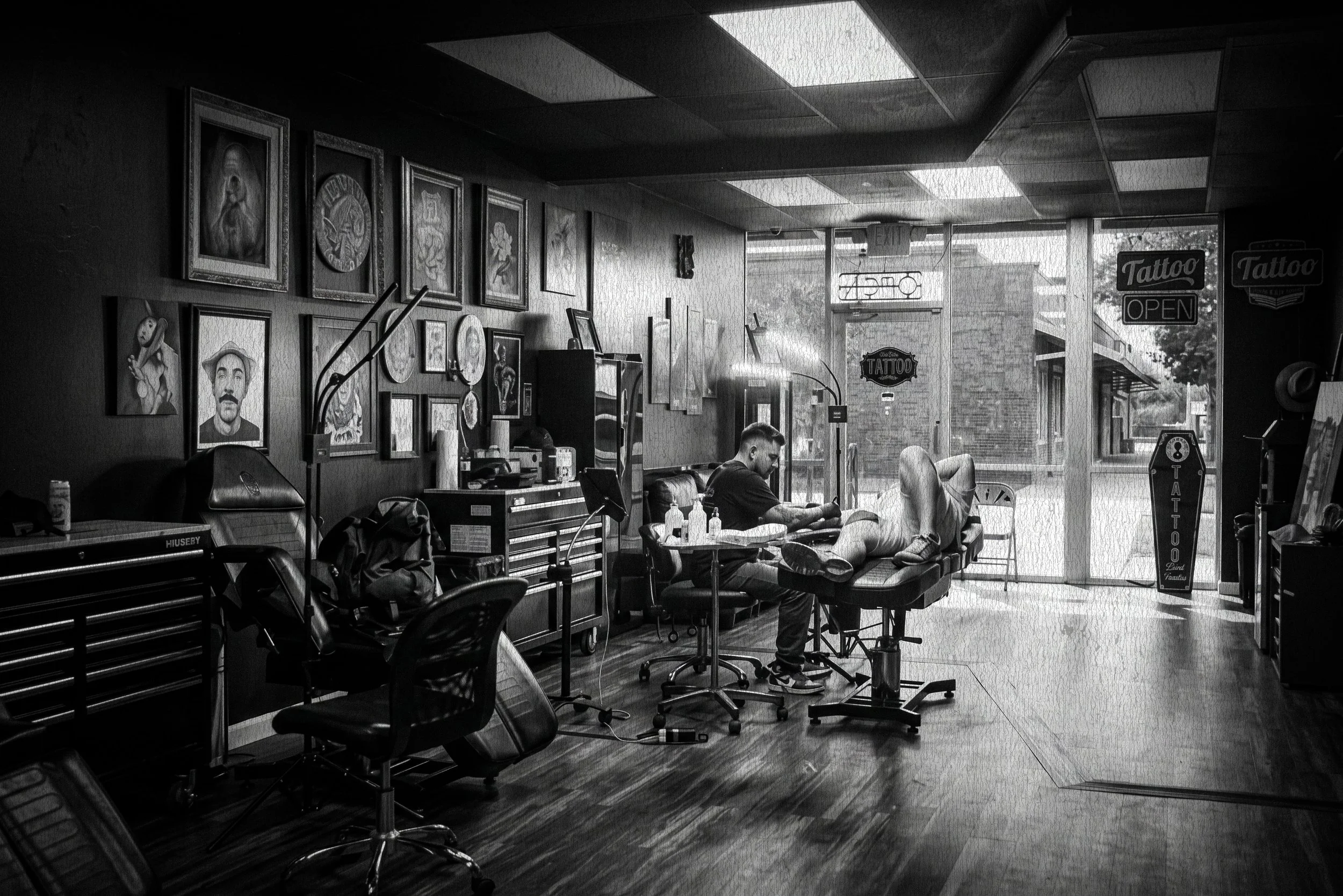 A tattoo shop with framed artwork on the wall, a waiting area with chairs, and a person receiving a tattoo while sitting in a chair. Signs for 'Tattoo' and 'Open' are visible outside the window.