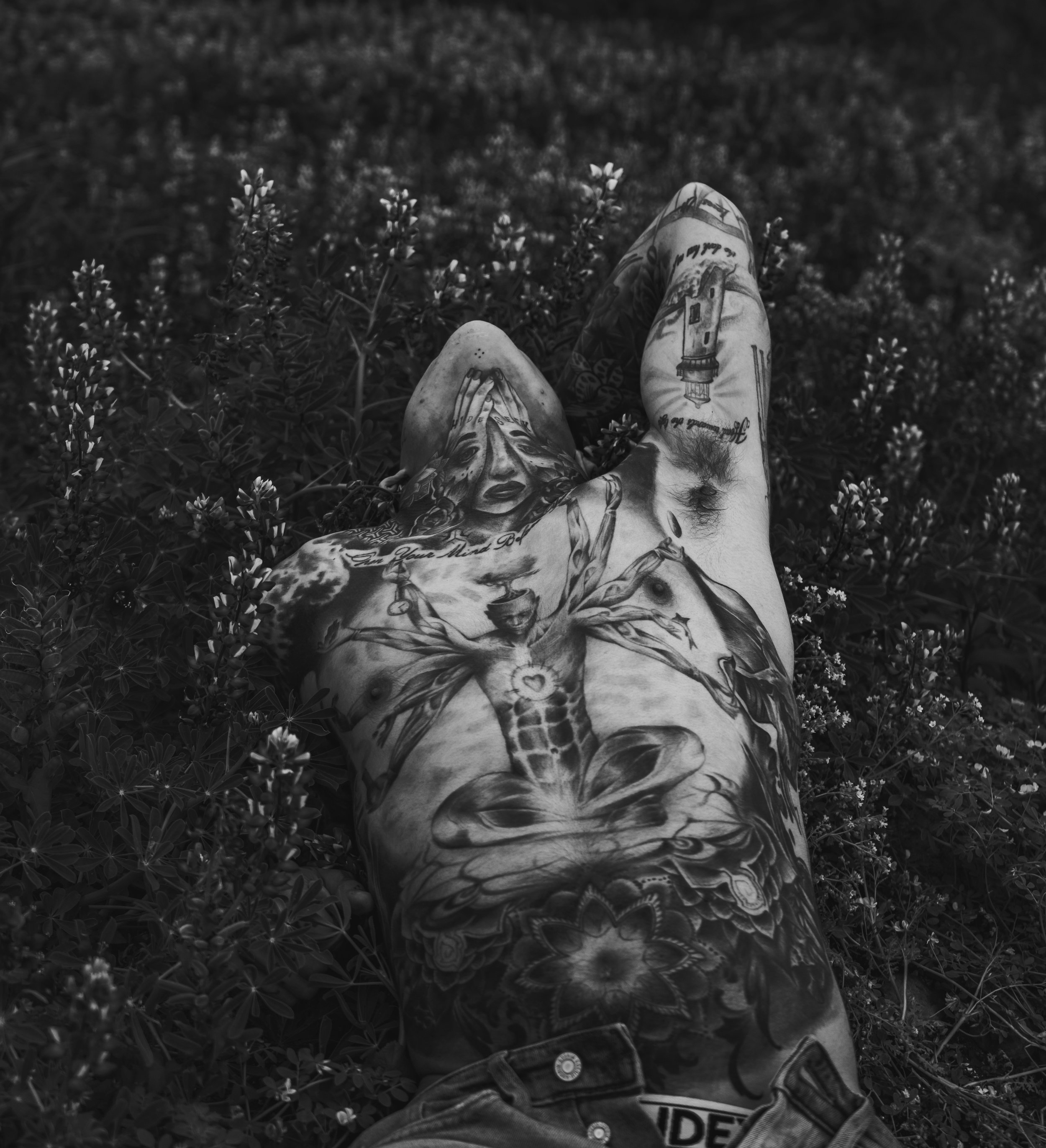 A person's tattooed legs and feet resting on a bed of small flowers, with tattoos of a woman's face, a lighthouse, and various designs visible.