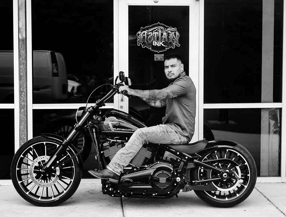 A man with tattoos sitting on a black Harley Davidson motorcycle in front of a glass storefront.
