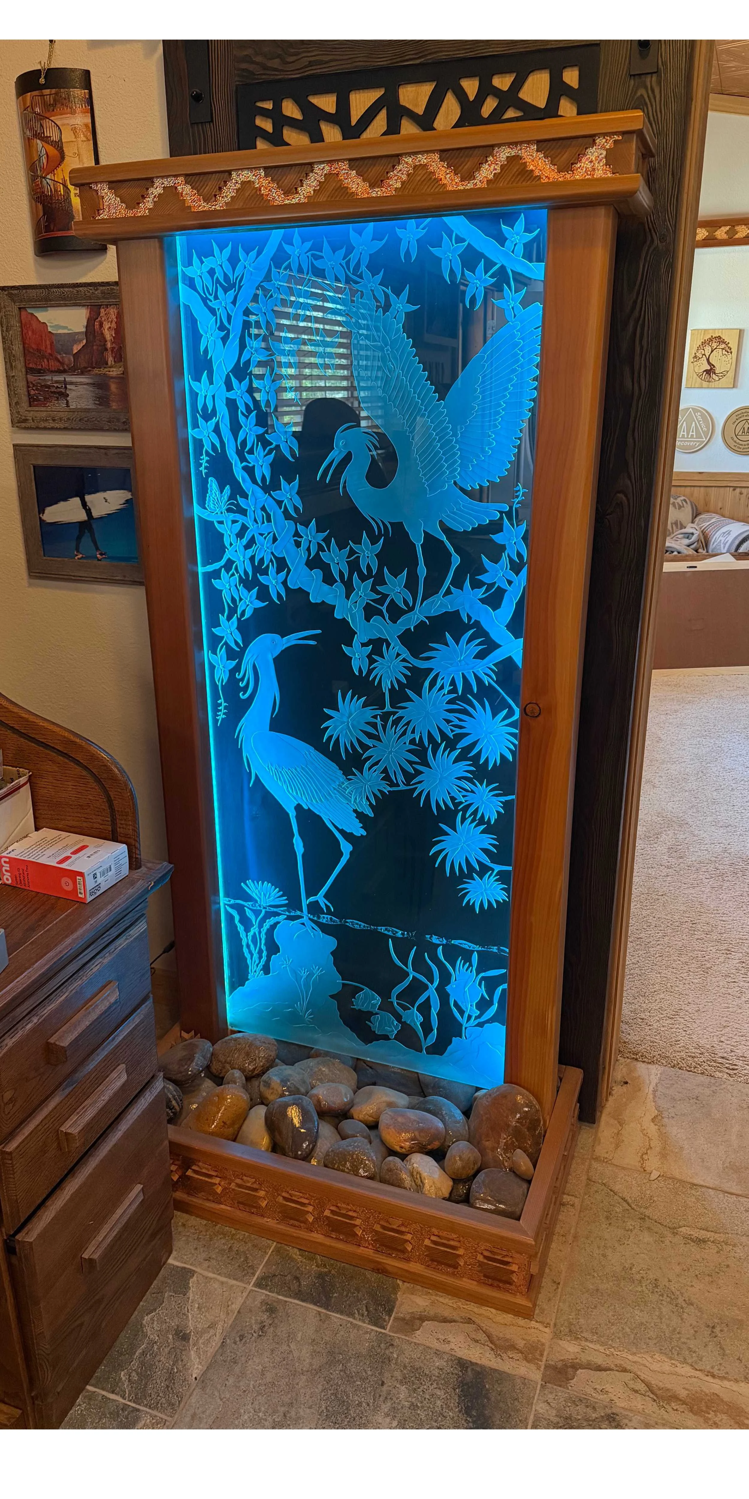 Decorative wooden illuminated water feature with a carved scene of two herons among foliage, illuminated with blue lighting, with stones at the base.