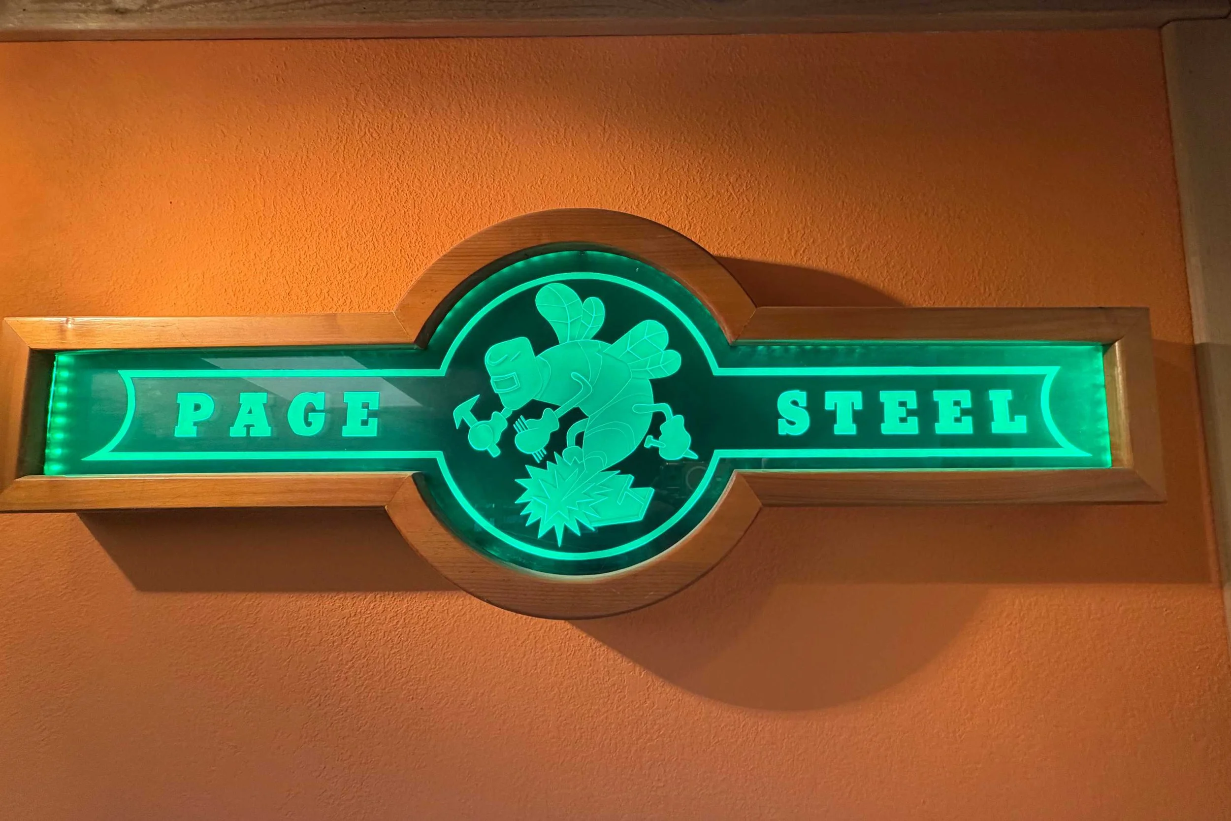 Neon sign with the words "Page Steel" and a cartoon bee holding a hammer, mounted on a wooden plaque on an orange wall.