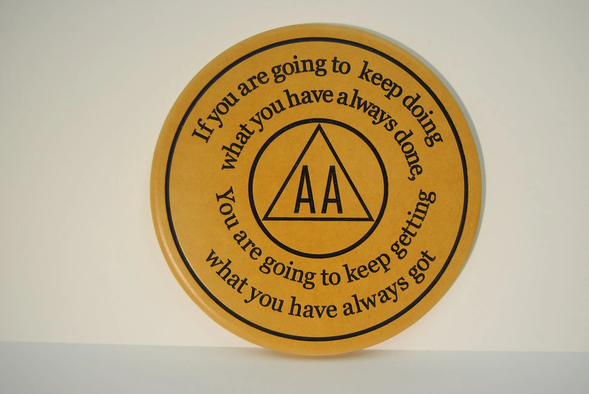 Orange circular plaque with black text and a black triangle logo with 'AA' inside. The text reads: 'If you are going to keep doing what you have always done, you will get what you always got.'