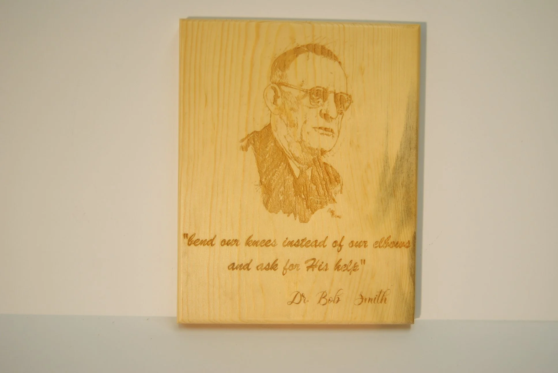 A wooden plaque with a sketch of a man wearing glasses and a graduation gown. The plaque has an inscription that reads, "bend our knees instead of our elbows and ask for His help" and is signed by Dr. Bob Smith.