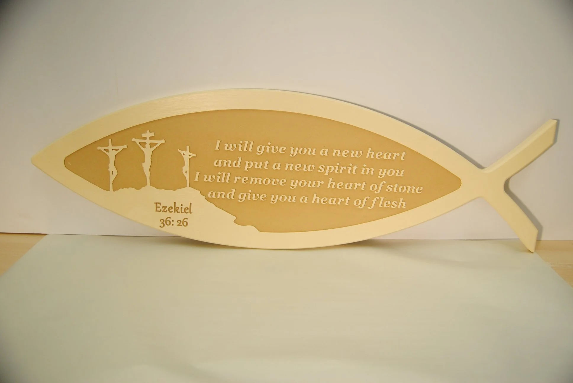 A fish-shaped wooden plaque with an engraving of three crosses and a scripture from Ezekiel 36:26 that reads, "I will give you a new heart and put a new spirit in you. I will remove your heart of stone and give you a heart of flesh."