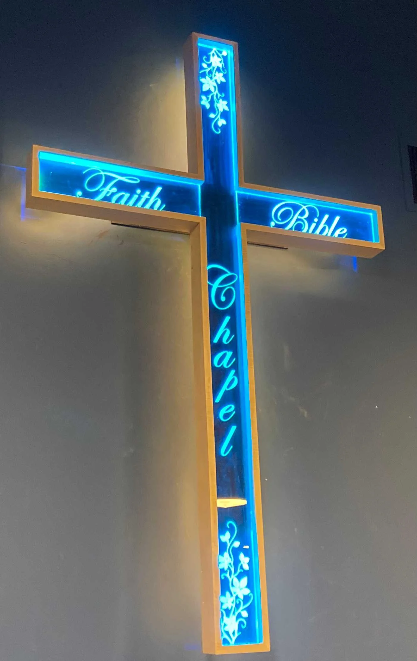 A vertical cross-shaped neon sign with blue lighting and decorative floral patterns. The sign has the words 'Faith', 'Hope', and 'Charity' written horizontally and vertically.