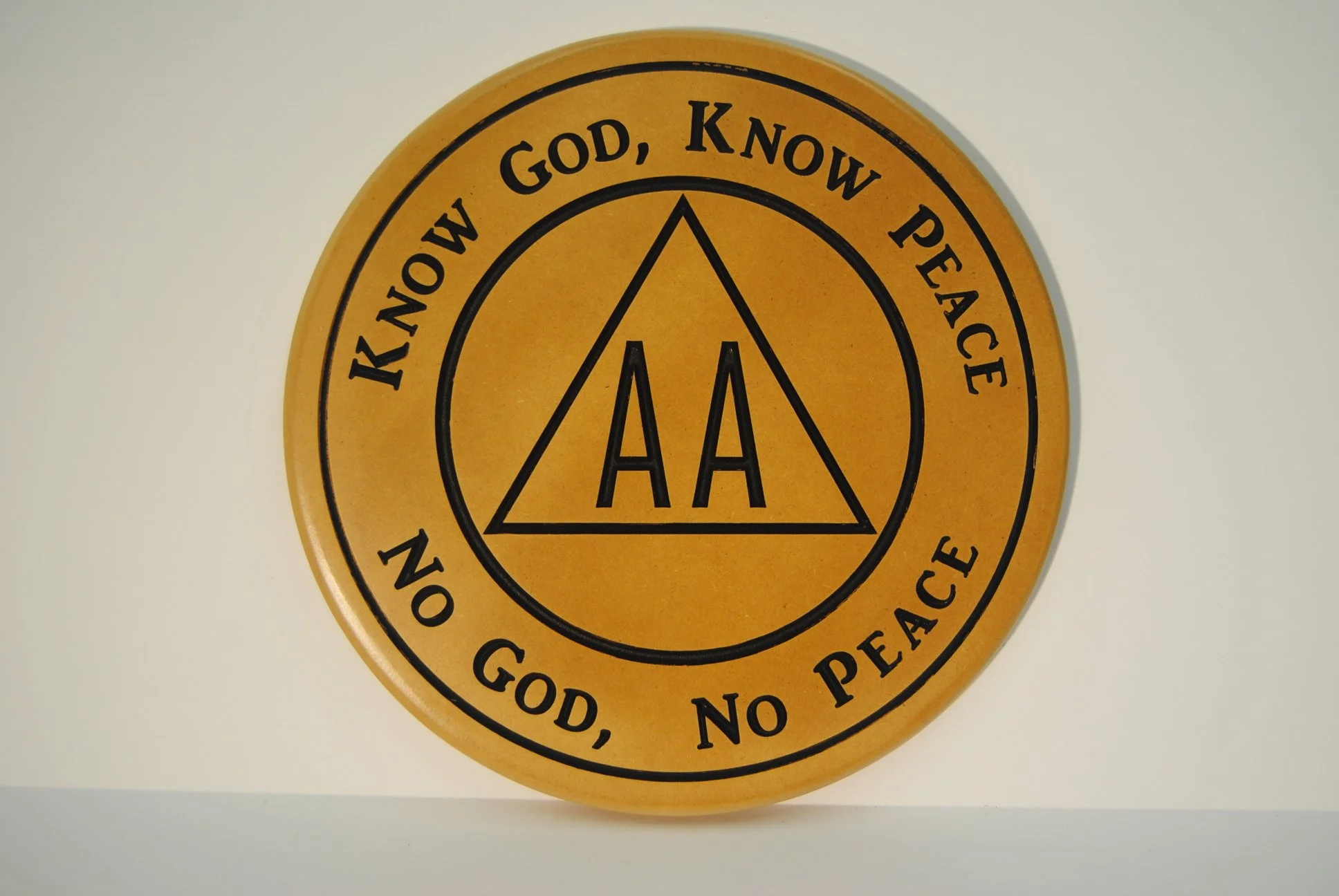 A circular yellow plaque with the words "Know God, Know Peace" and "No God, No Peace" surrounding the AA logo, which is an uppercase letter A within a triangle.