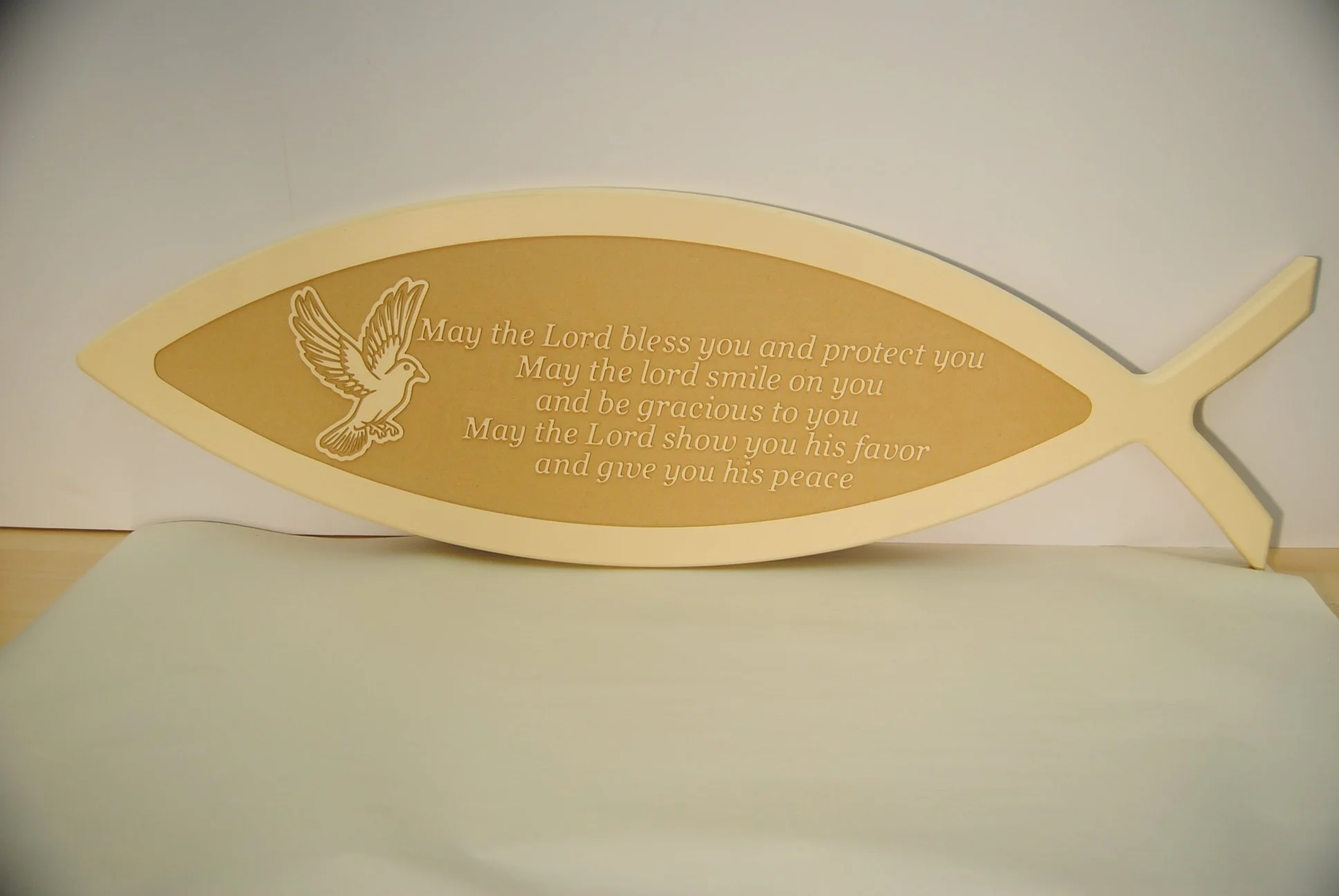 A fish-shaped wooden plaque with gold coloring, engraved with a religious prayer and a dove illustration.