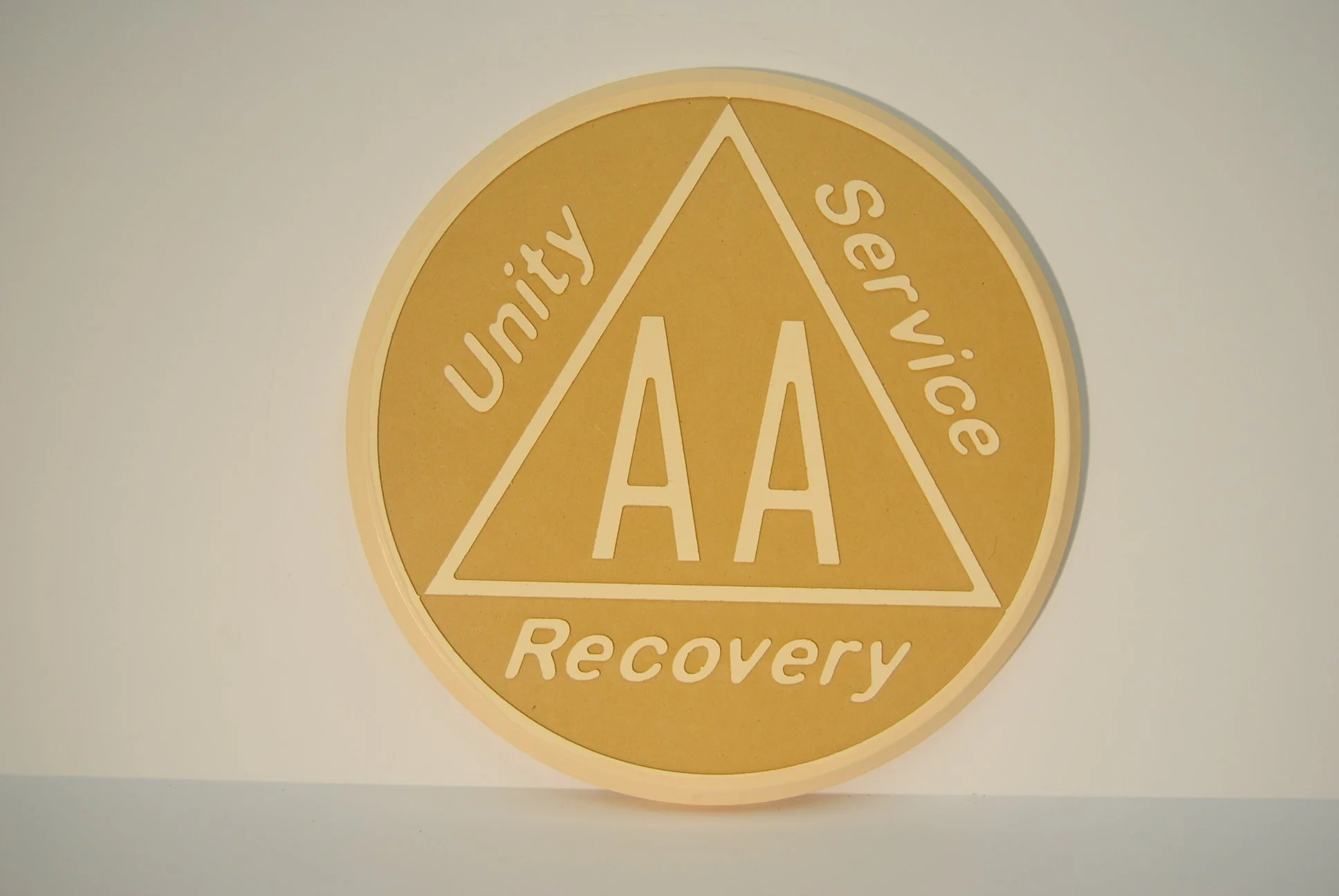 A circular plaque with a gold background, featuring a triangle and the words 'Unity', 'Service', and 'Recovery' around it, and the letter 'A' in the center.