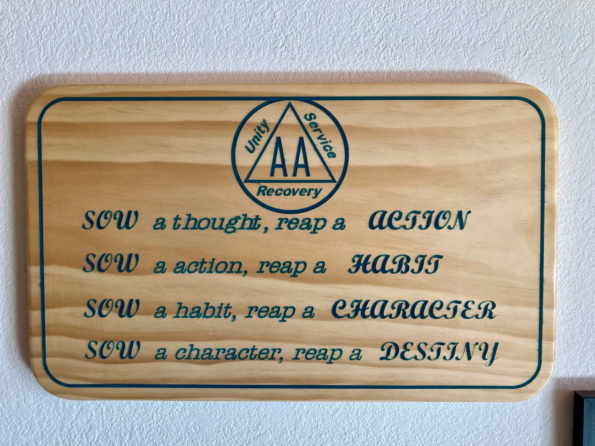 Wooden plaque with blue text and a circle logo featuring a triangle with the letters A A and the words unity, service, and recovery on a white textured wall.