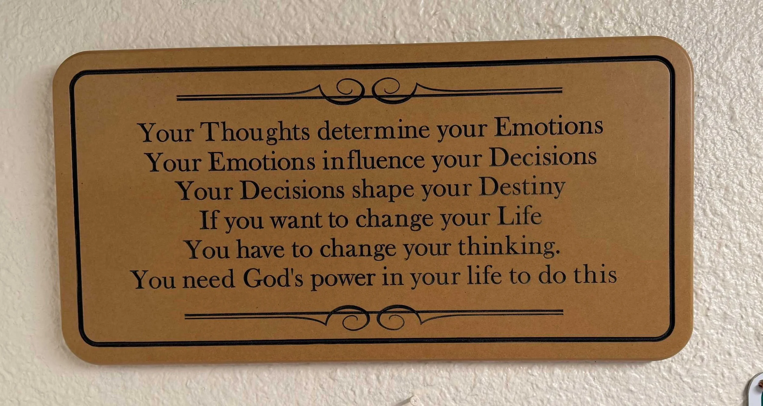 A motivational quote on a tan plaque with black border and decorative swirls, affixed to a textured white wall.