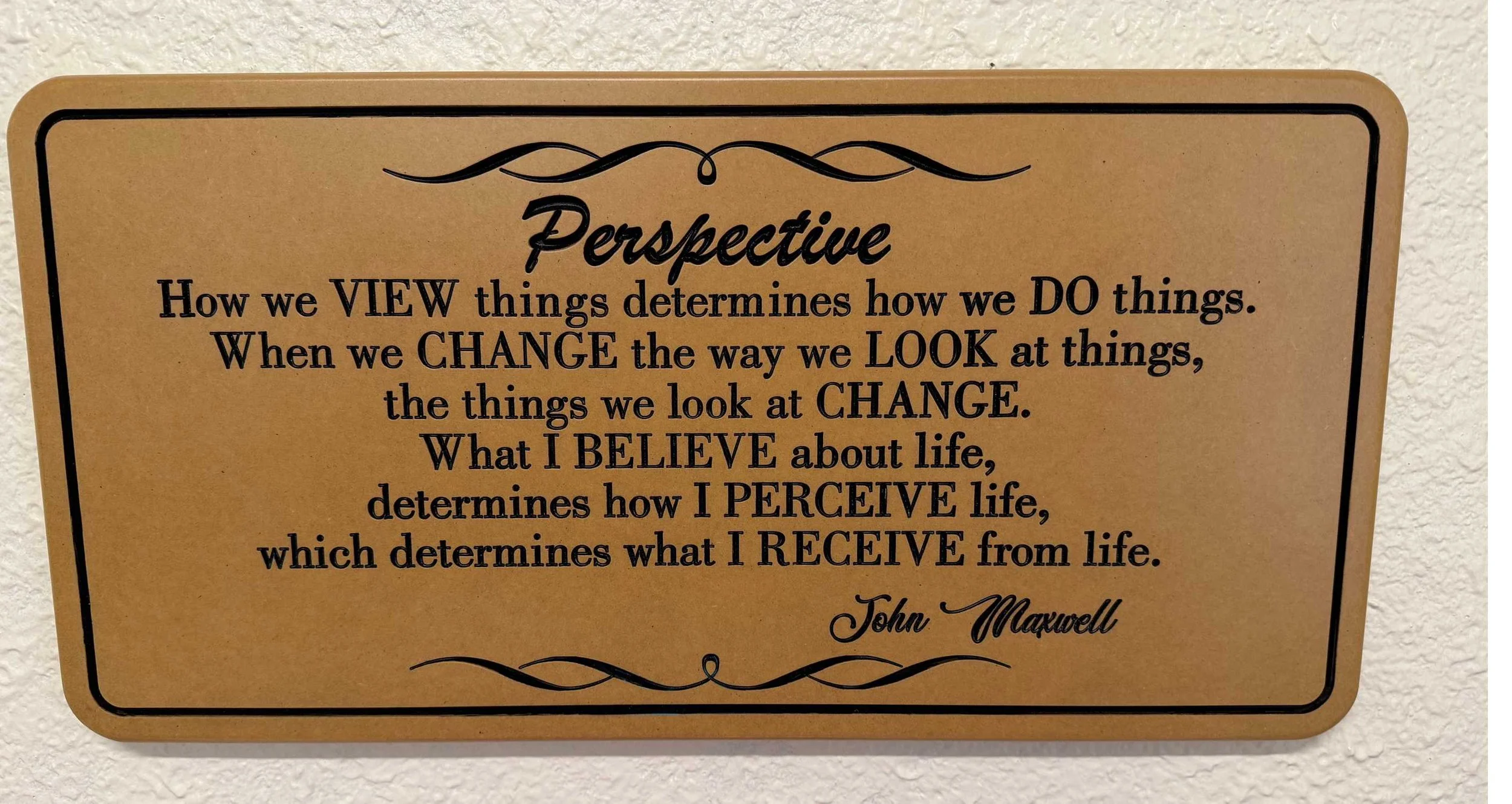 A motivational quote on a beige plaque mounted on a wall. The quote discusses perspective, change, and perception in life, attributed to John Maxwell.