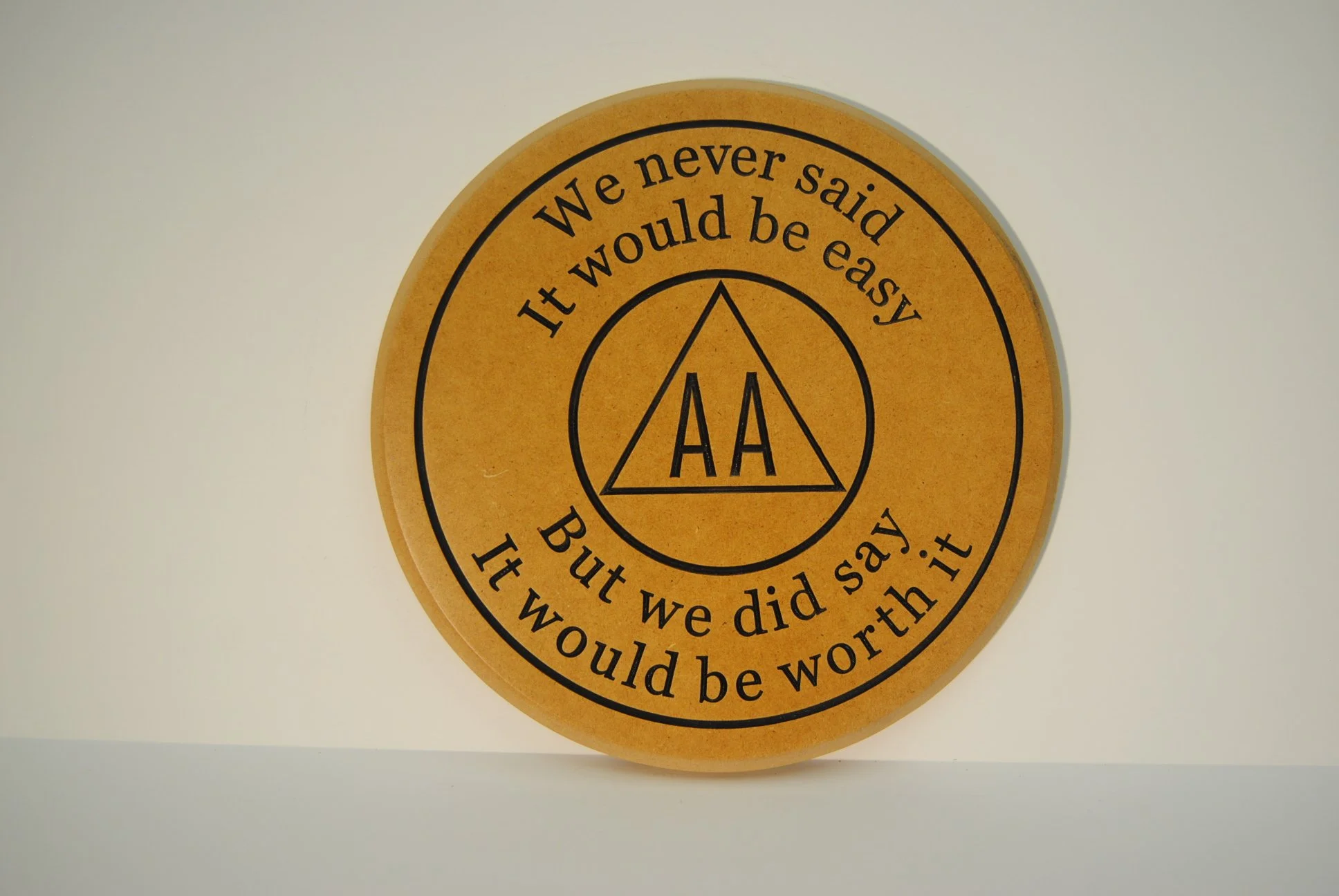 A circular orange plaque with black text and the AA logo in the center. The text reads: 'We never said it would be easy, it would be worth it. AA.'