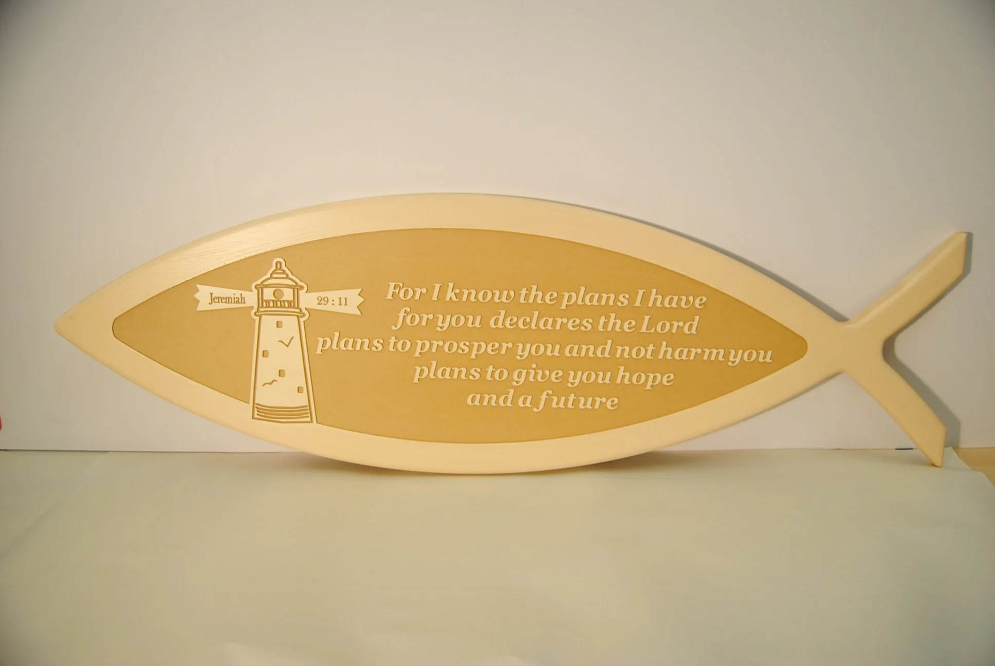 A fish-shaped wooden plaque with a gold background features a quote from Jeremiah 29:11 about God's plans to prosper and give hope and a future. The design includes a lighthouse illustration and a banner with the scripture reference.