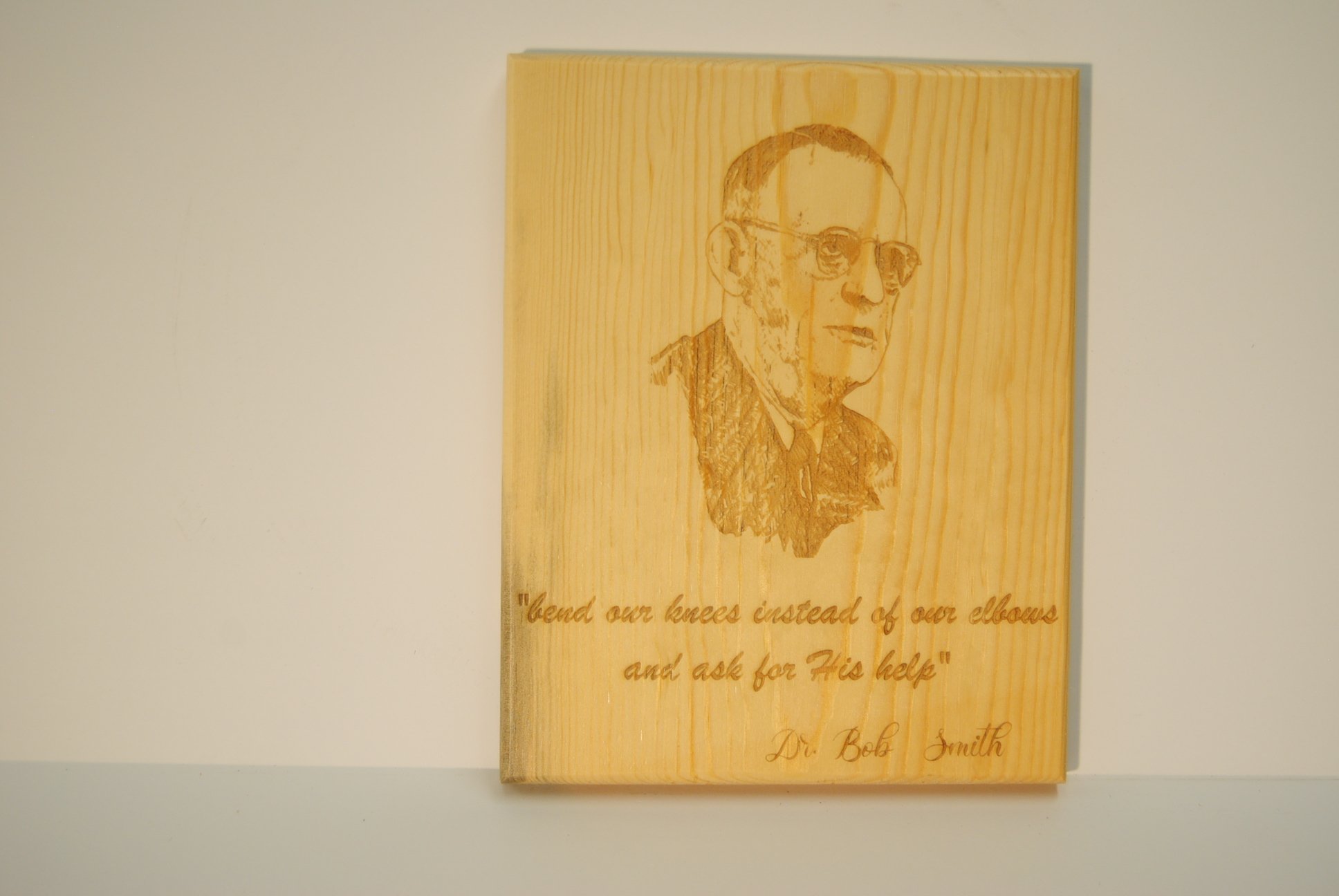 A wooden plaque with a carved portrait of Dr. Bob Smith, featuring glasses and a serious expression, and an engraved quote that reads, 'bend our knees instead of our elbows and ask for His help'.