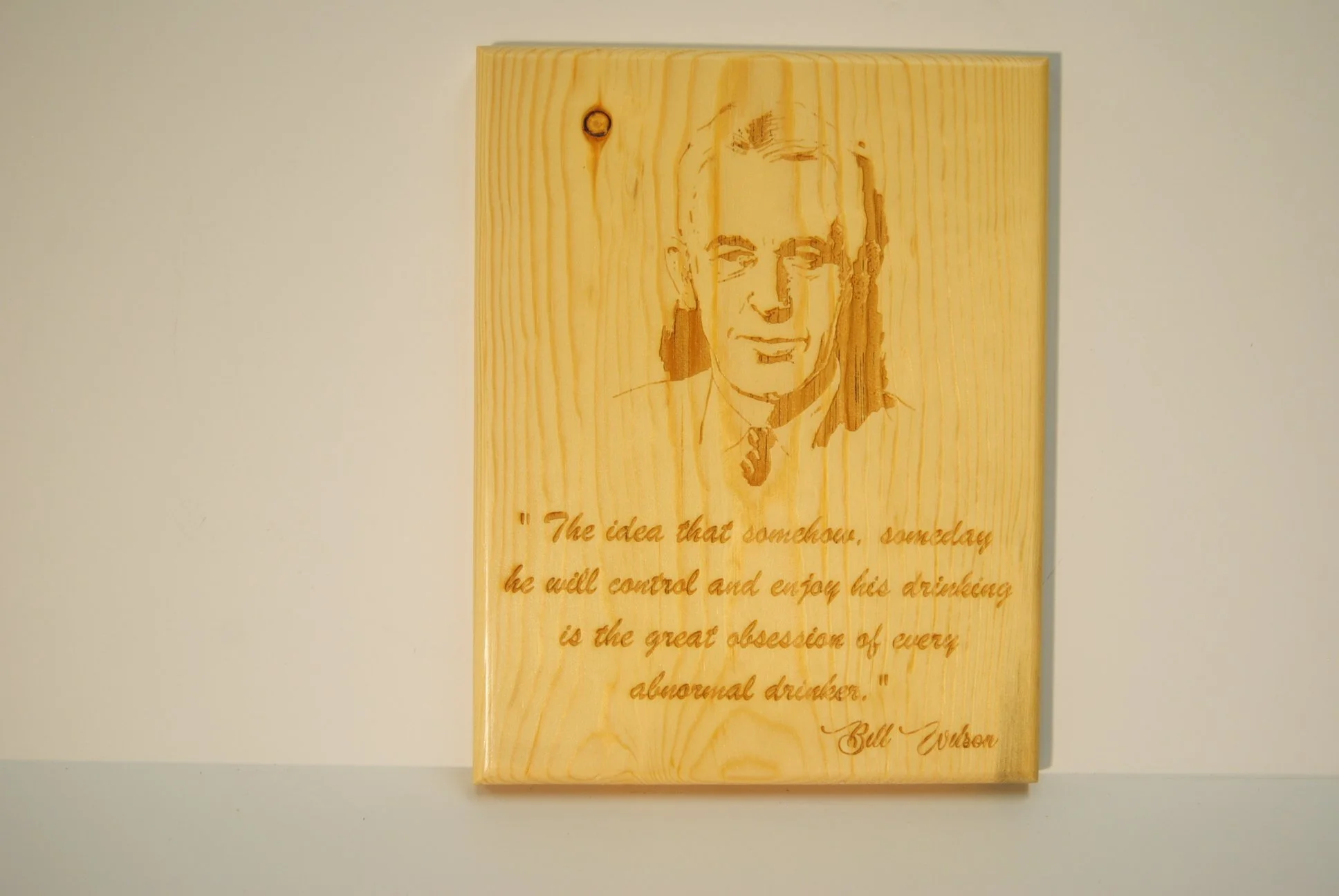 A wooden plaque with a portrait burned into it of a man with short hair and a slight smile. Below the portrait, there is an inscription that reads: 'The idea that someday he will control and enjoy his drinking is the great obsession of every alcoholi