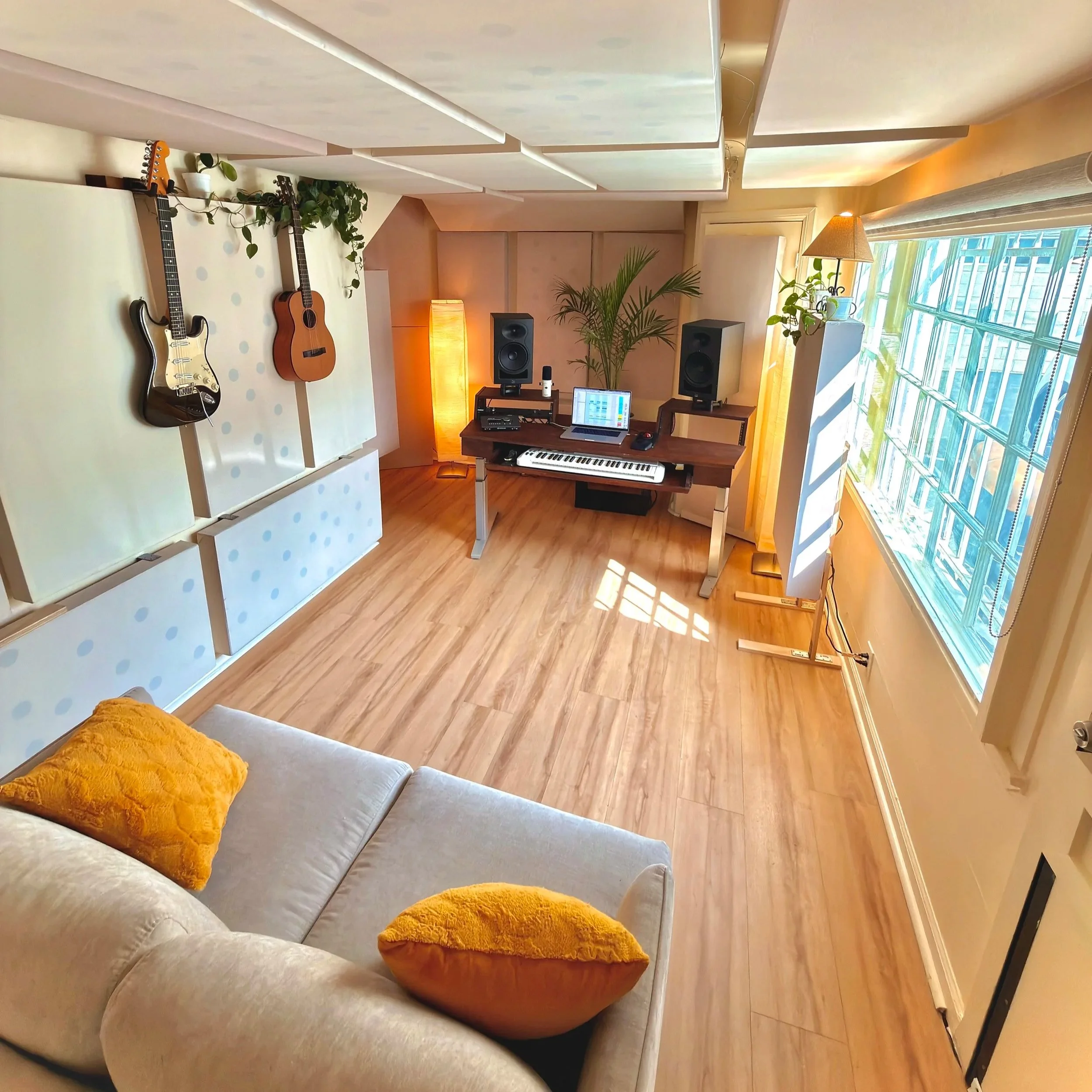 A cozy living room with a gray sofa decorated with two orange cushions, a white table, a music workstation with two guitars, a keyboard, laptop, speakers, and a small plant by a large window with curtains and blinds.