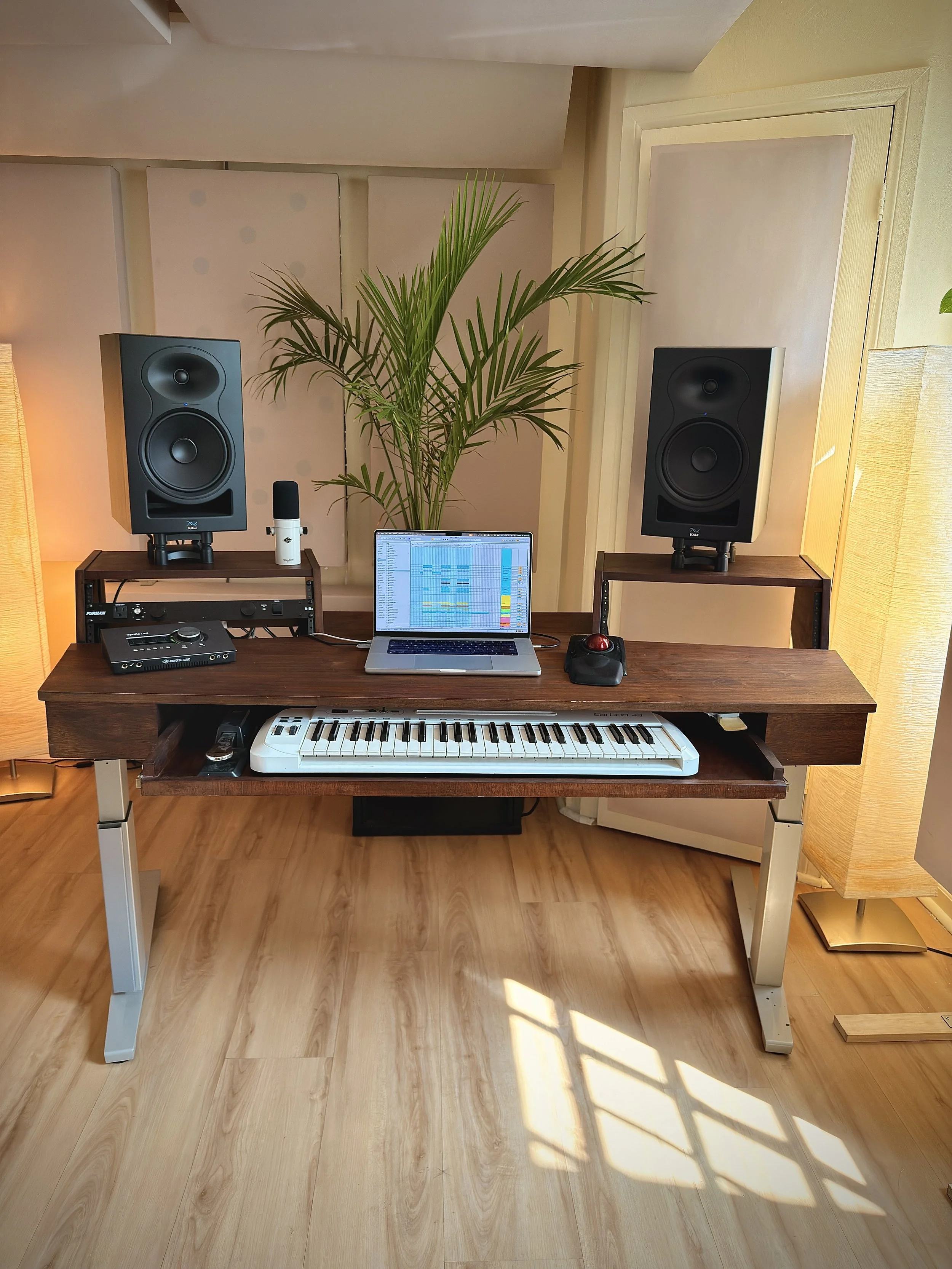 Music production workspace featuring a wooden desk with a MIDI keyboard, two large black studio monitor speakers on stands, a laptop displaying digital audio workstation software, a microphone, a mouse, and a device connected to the laptop. A tall green plant is behind the workspace, with a white wall and acoustic panels in the background, and warm yellow lighting.
