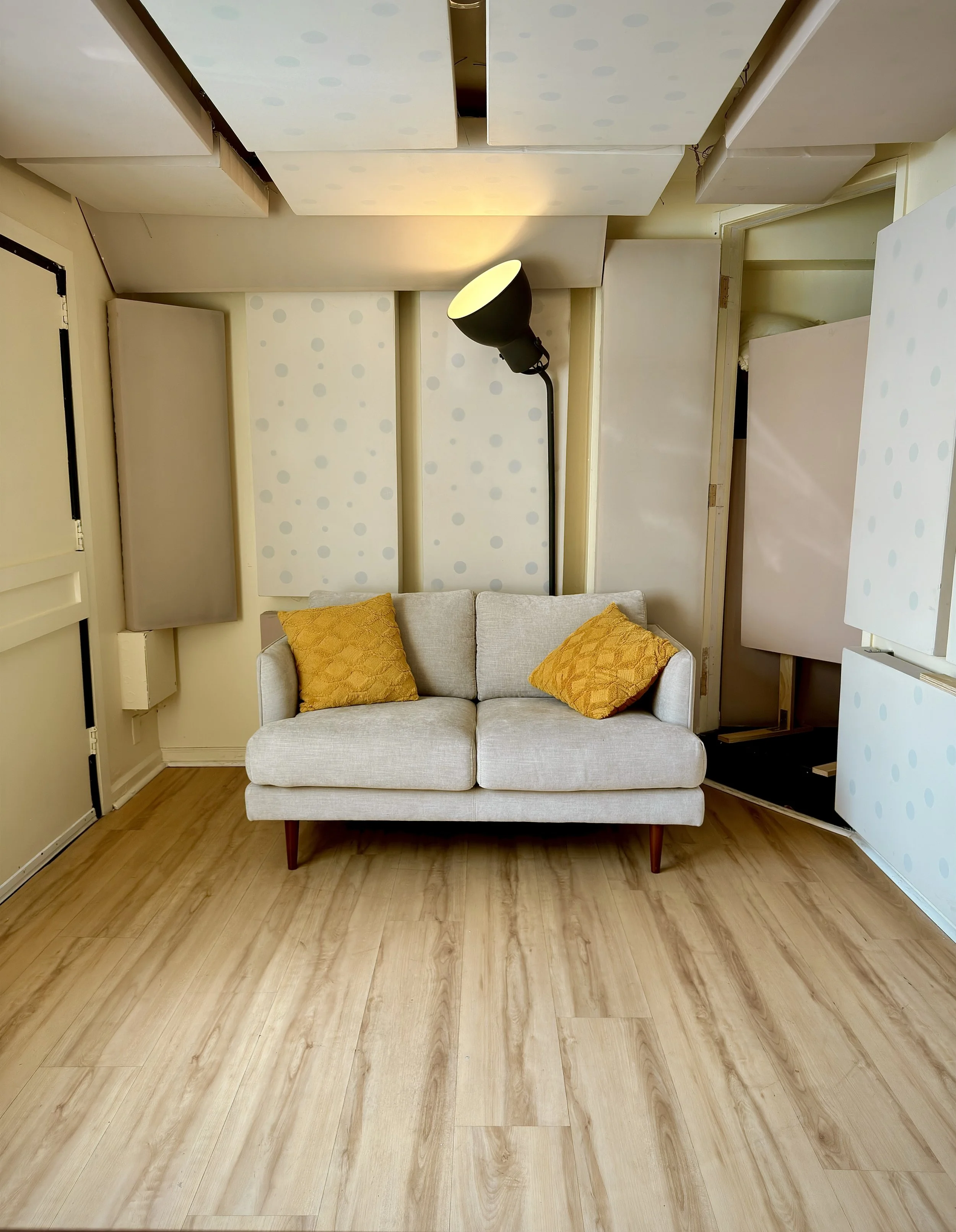 A beige sofa with two yellow cushions sits against a wall with partially installed decorative panels. A black floor lamp illuminates the wall, and a wooden floor extends across the room.
